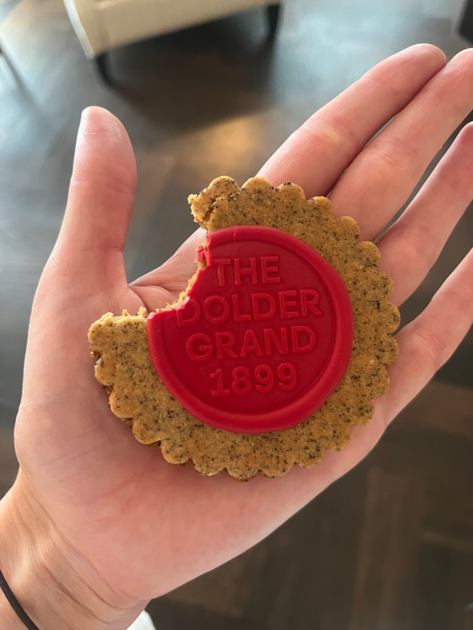 A hand holds a round biscuit with a decorative red icing centre that reads THE DOLDER GRAND 1899. The biscuit, inspired by the fairytale hotel in Zurich, has a bite taken out of it.