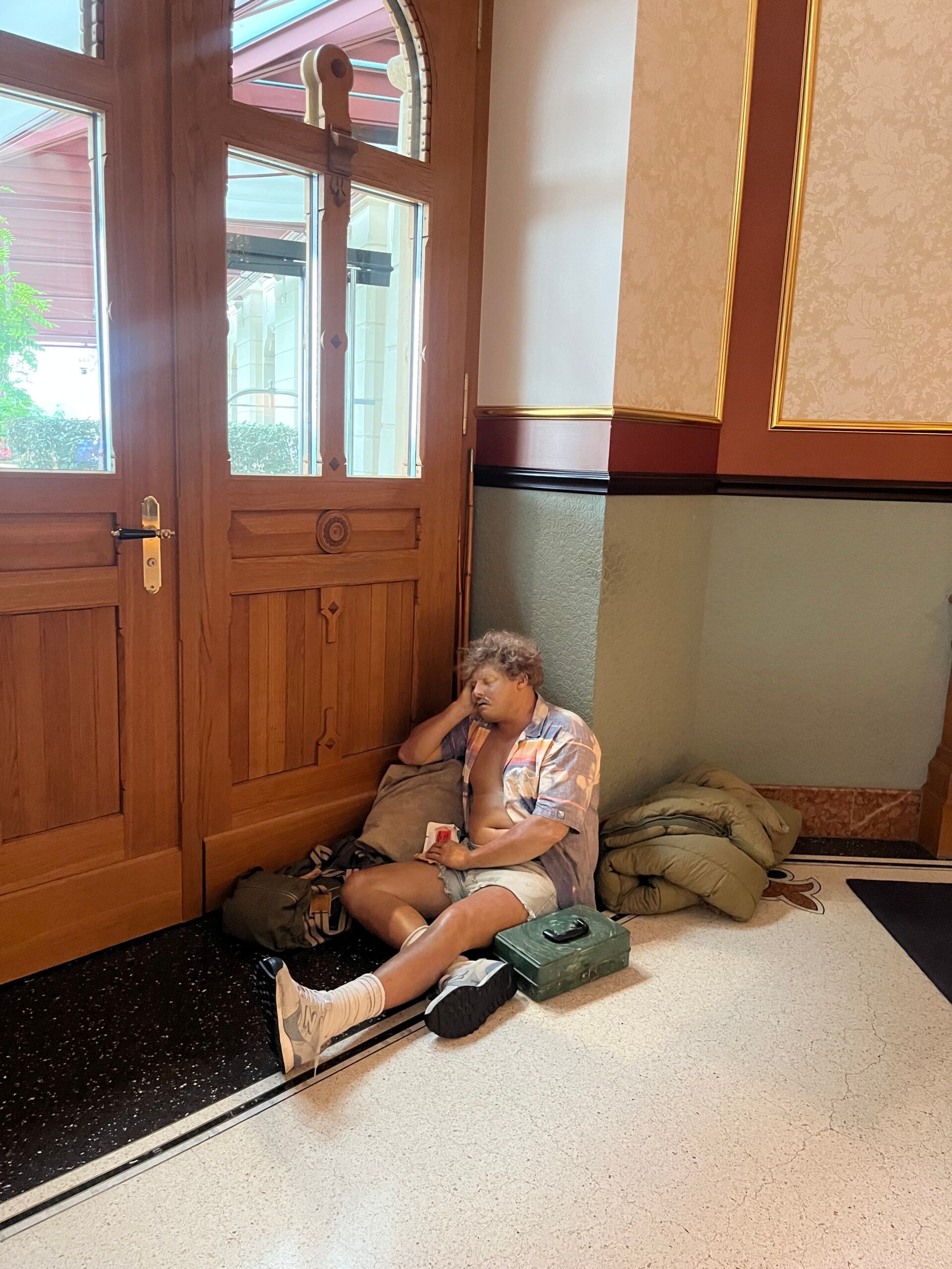 A person with curly hair, wearing a light shirt, shorts, and white trainers, sits on the floor inside a building near wooden doors at the Dolder Grand in Zurich, with bags and belongings placed around them, evoking a scene from a fairy-tale hotel.