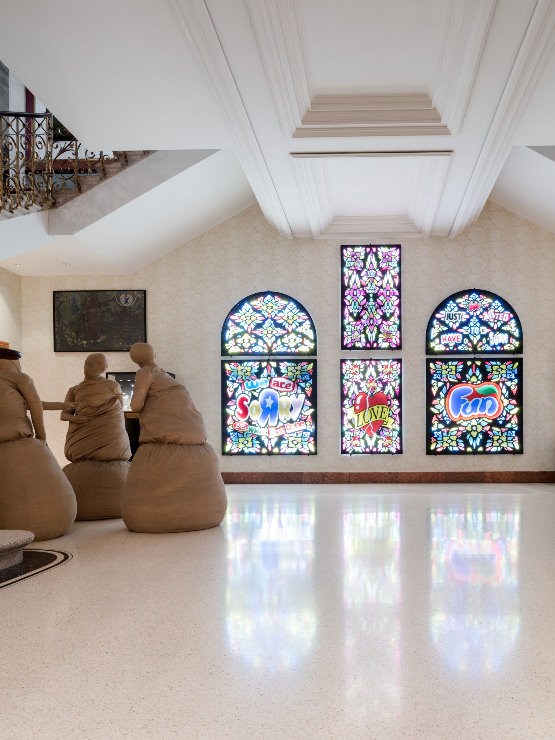 A bright room at the fairytale hotel Dolder Grand in Zurich features stained glass windows with the words Soul, Love, and Fun. Four abstract human sculptures sit at a curved bar, whilst a staircase with an ornate railing appears in the background.