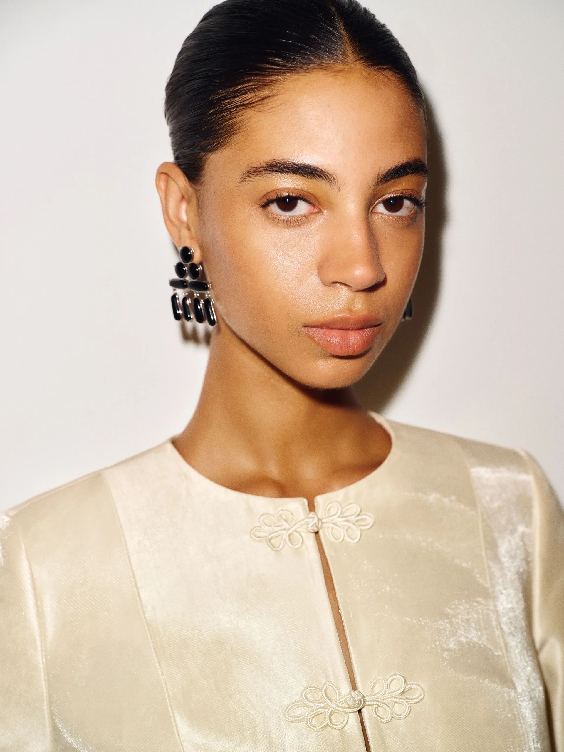 Woman with slicked-back hair wears a cream-coloured, textured jacket with floral fastenings and large black earrings, showcasing an elegant fashion detail. She looks directly at the camera with a neutral expression against a plain light background.