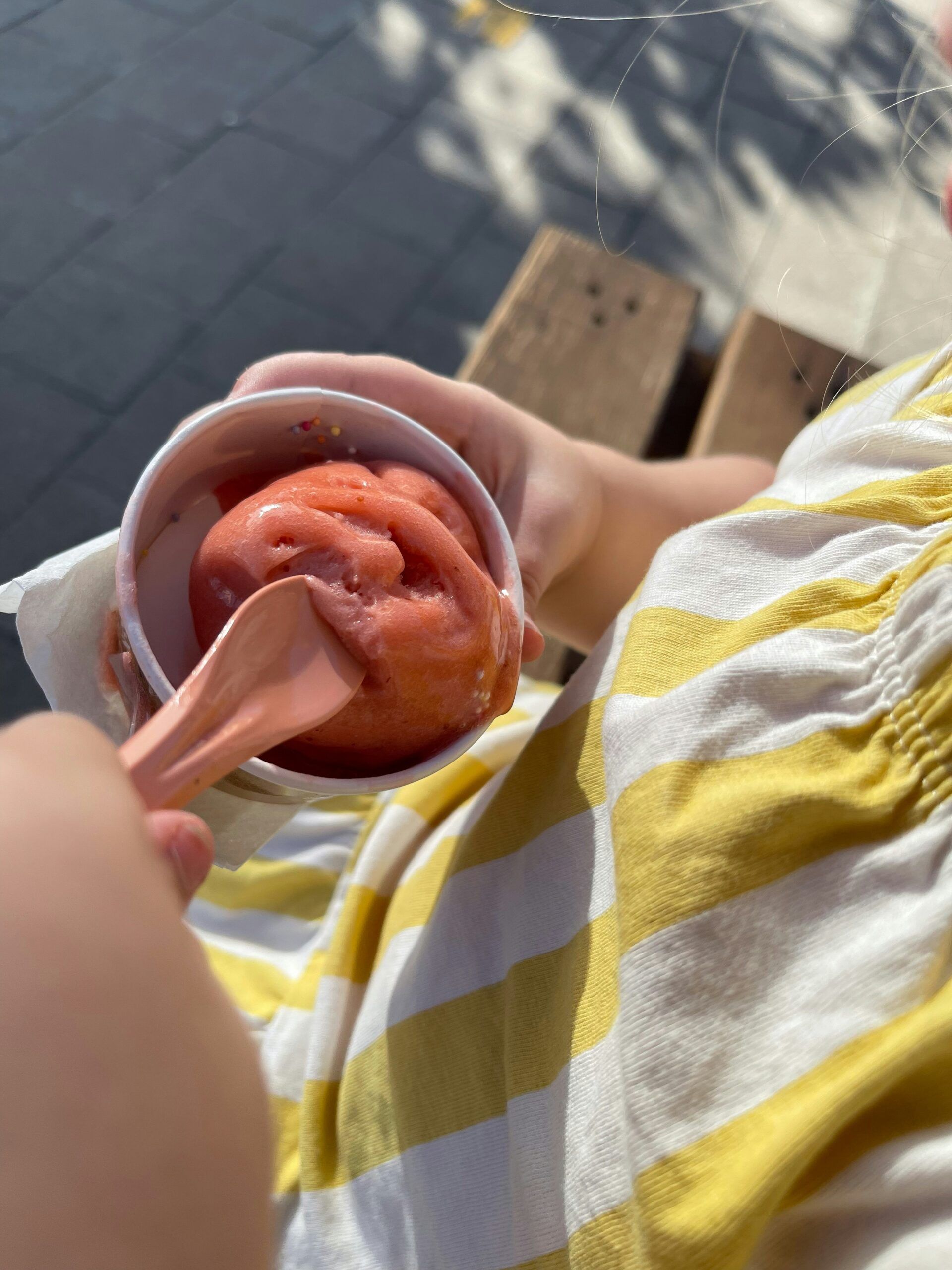A child in a yellow and white striped shirt holds a cup of red sorbet and eats it with a pink spoon whilst sitting on a bench outdoors, enjoying the simple pleasures of Summer Holidays With Kids.