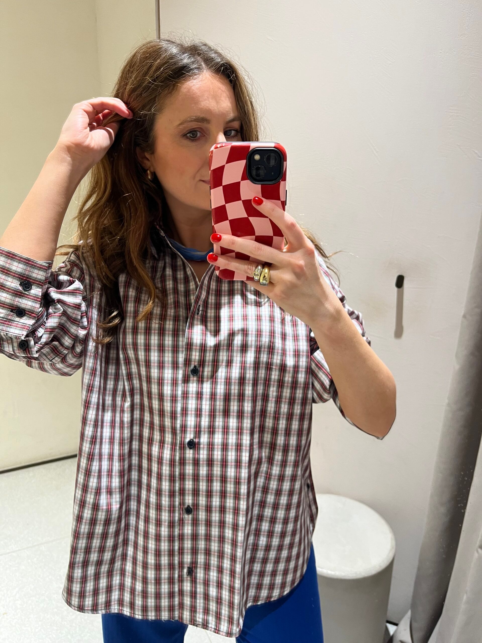 A woman with wavy hair takes a mirror selfie wearing a checked shirt, blue trousers, and holding a red and white chequered phone case—her effortlessly chic look showcases pieces that are new in at Zara. She stands in a bright, minimal room with white walls and a round seat.