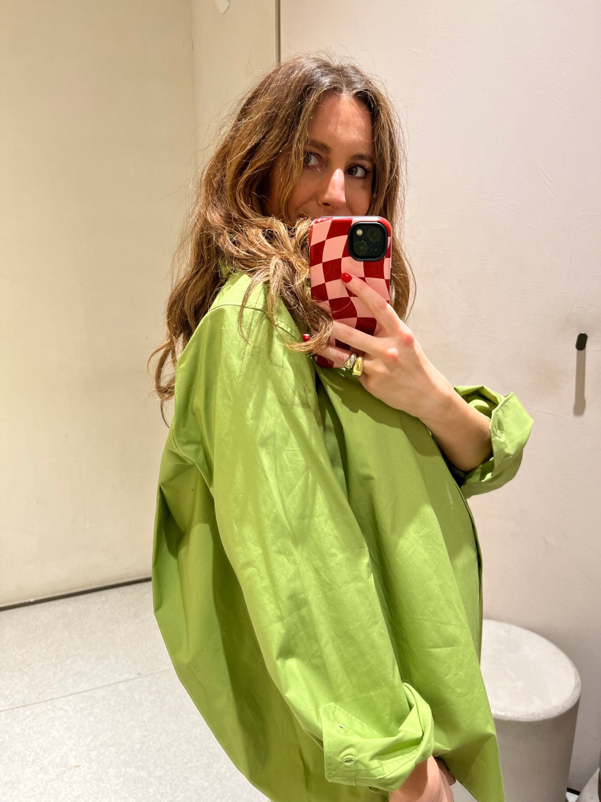 Woman with long brown hair wearing a loose, bright green shirt—new in at Zara—takes a mirror selfie with a red and white chequered phone case in a minimal room.