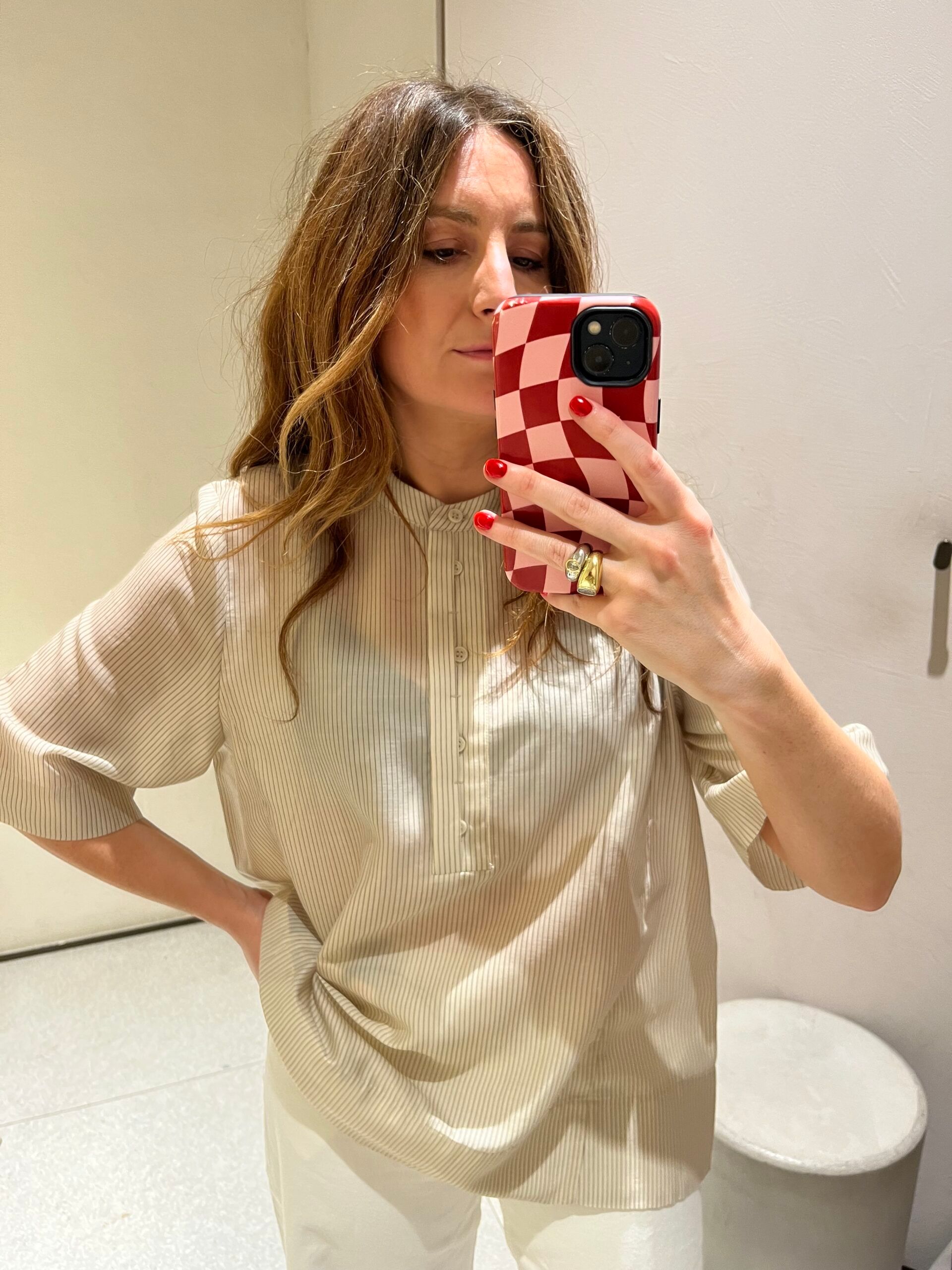 Woman taking a mirror selfie, wearing a light beige blouse and white trousers—both new in at Zara. Her phone has a red and white chequered case, and she has wavy brown hair. The background is a minimal indoor space.