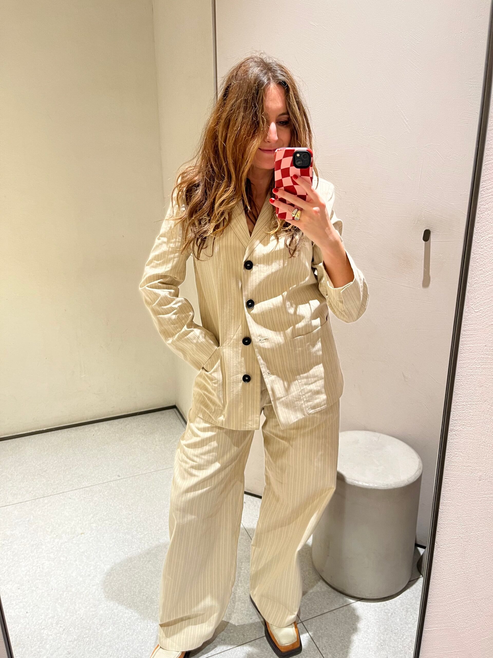 Woman with long hair wears a cream pinstripe suit and white shoes, taking a mirror selfie in a minimal dressing room with neutral walls and a small round stool—her look is effortlessly chic and new in at Zara.