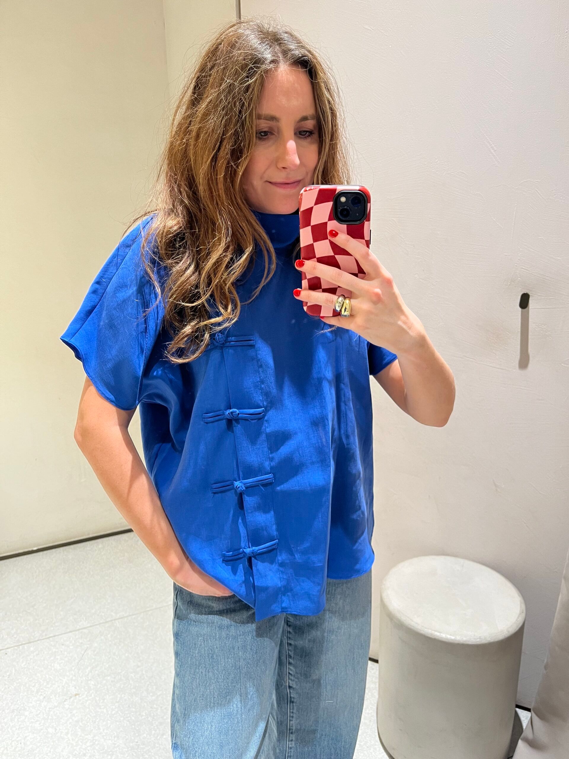 Woman with long brown hair wears a vibrant blue, short-sleeved top with knot details and light blue jeans—an effortlessly stylish look that’s new in at Zara. She takes a mirror selfie with a red and white chequered phone case, while a grey stool stands to her right.