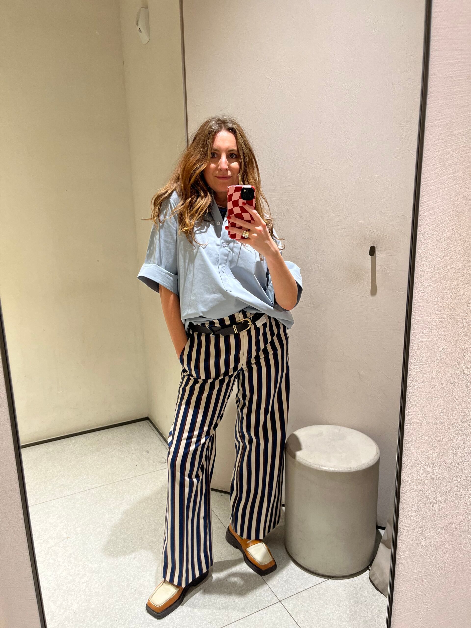 Woman taking a mirror selfie in a changing room, wearing a light blue shirt and high-waisted blue and white striped trousers, with brown and white shoes—both new in at Zara. She has one hand in her pocket.