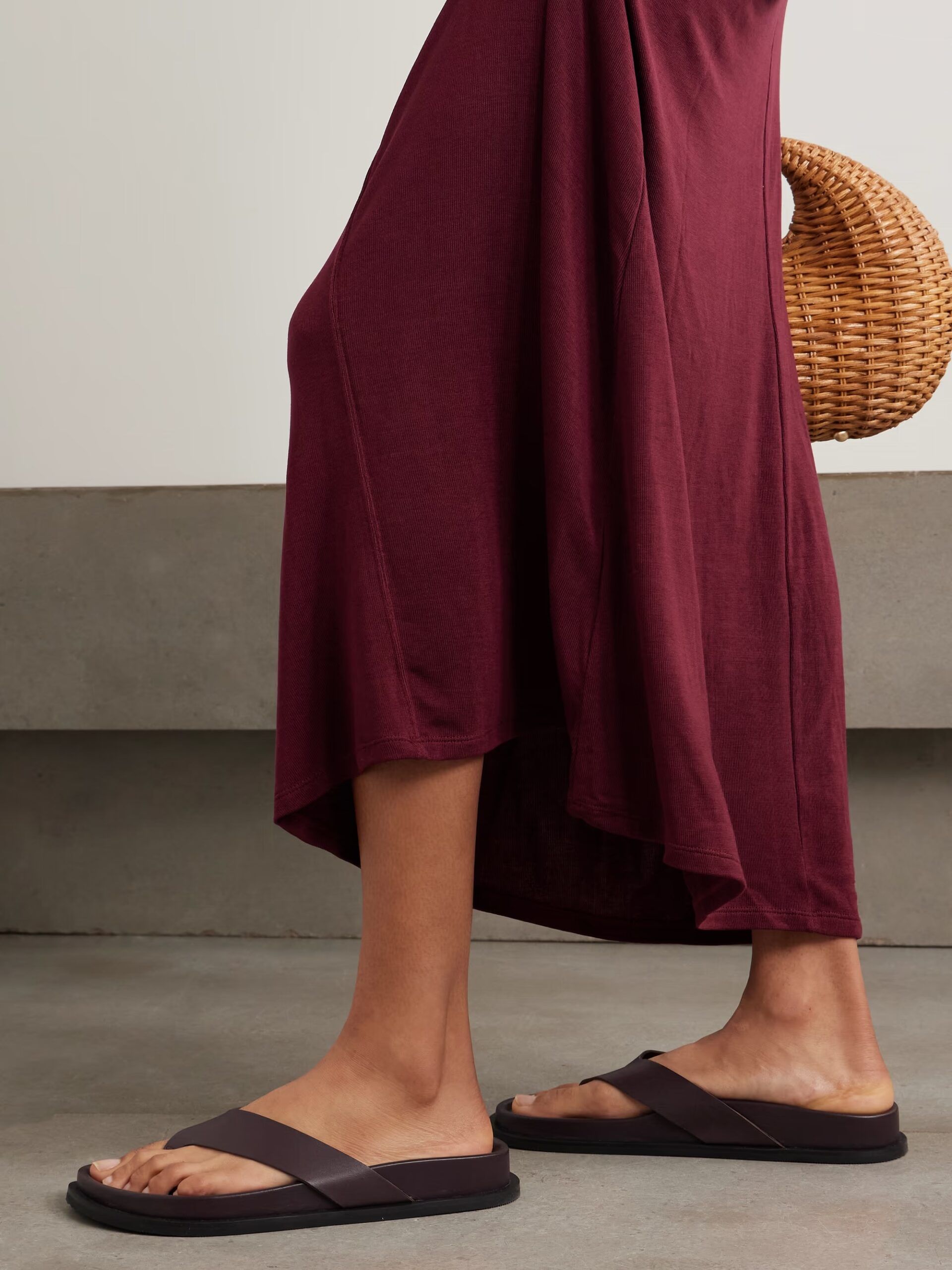 A person wearing a long burgundy skirt and dark sandals stands indoors, holding a round woven bag—an effortlessly chic look perfect for the summer sale season. Only the lower half of their body is visible.