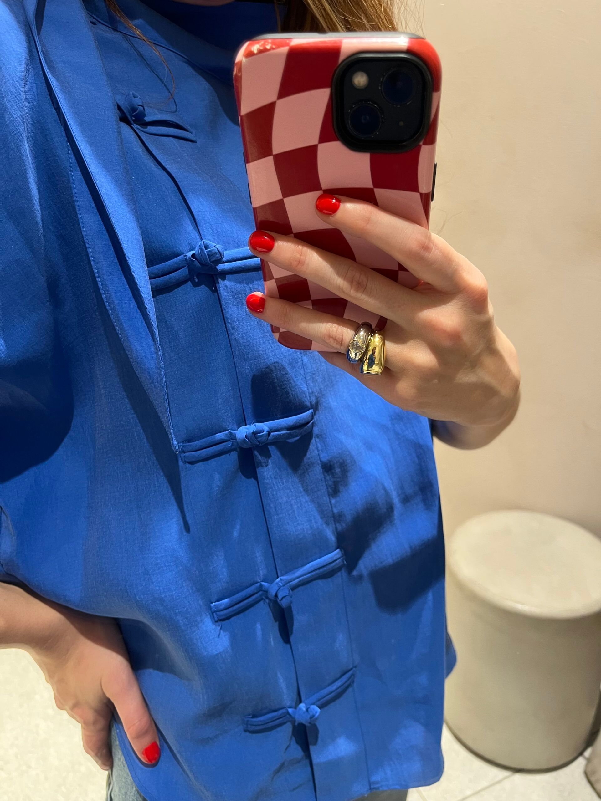 A person wearing a bright blue top reminiscent of International Klein Blue with knotted buttons takes a mirror selfie, holding a red chequered phone. They have red nail varnish and wear multiple rings on their hand, adding a pop of colour everywhere in the shot.