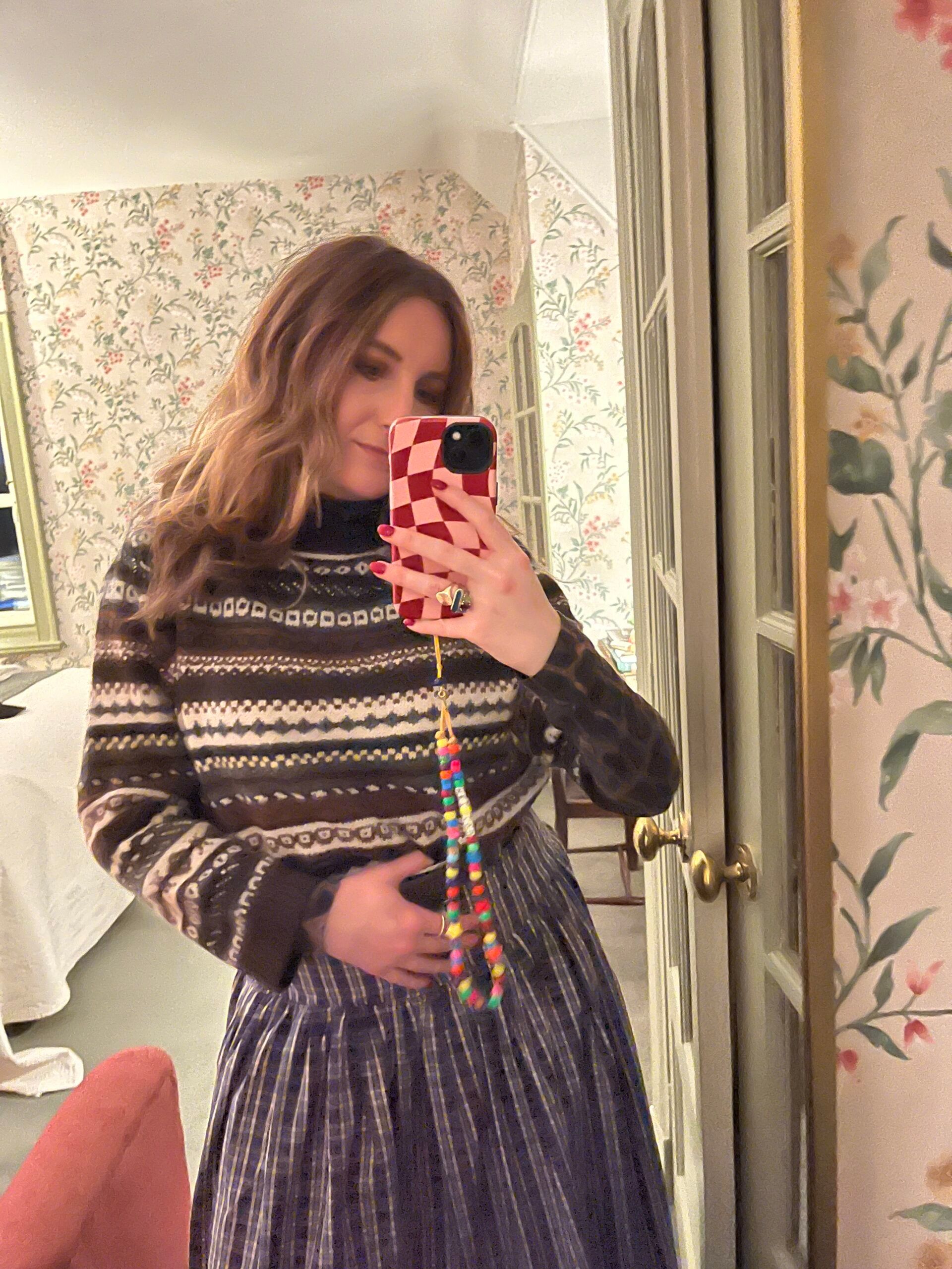 A woman with wavy hair takes a mirror selfie in a cosy, floral-wallpapered room. Showcasing her unique fashion sense, she wears a patterned jumper and striped skirt, holding a phone with a red chequered case and beaded strap.