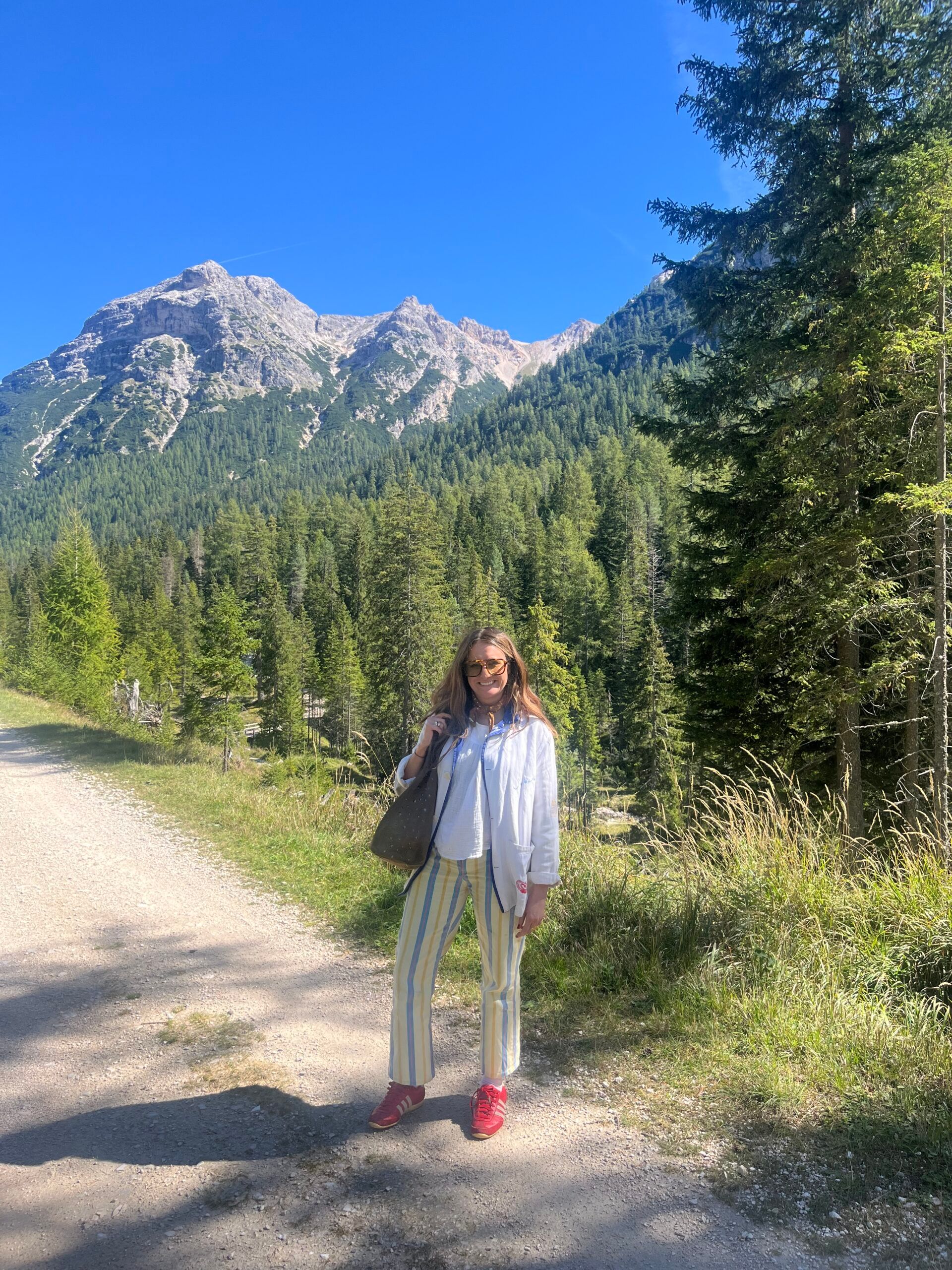 A woman stands on a sunny forest path with mountains in the background, confidently wearing the striped trousers and light shirt from the collection of clothes my boyfriend hates, along with red shoes and sunglasses. She is holding a bag, surrounded by tall trees and blue sky.