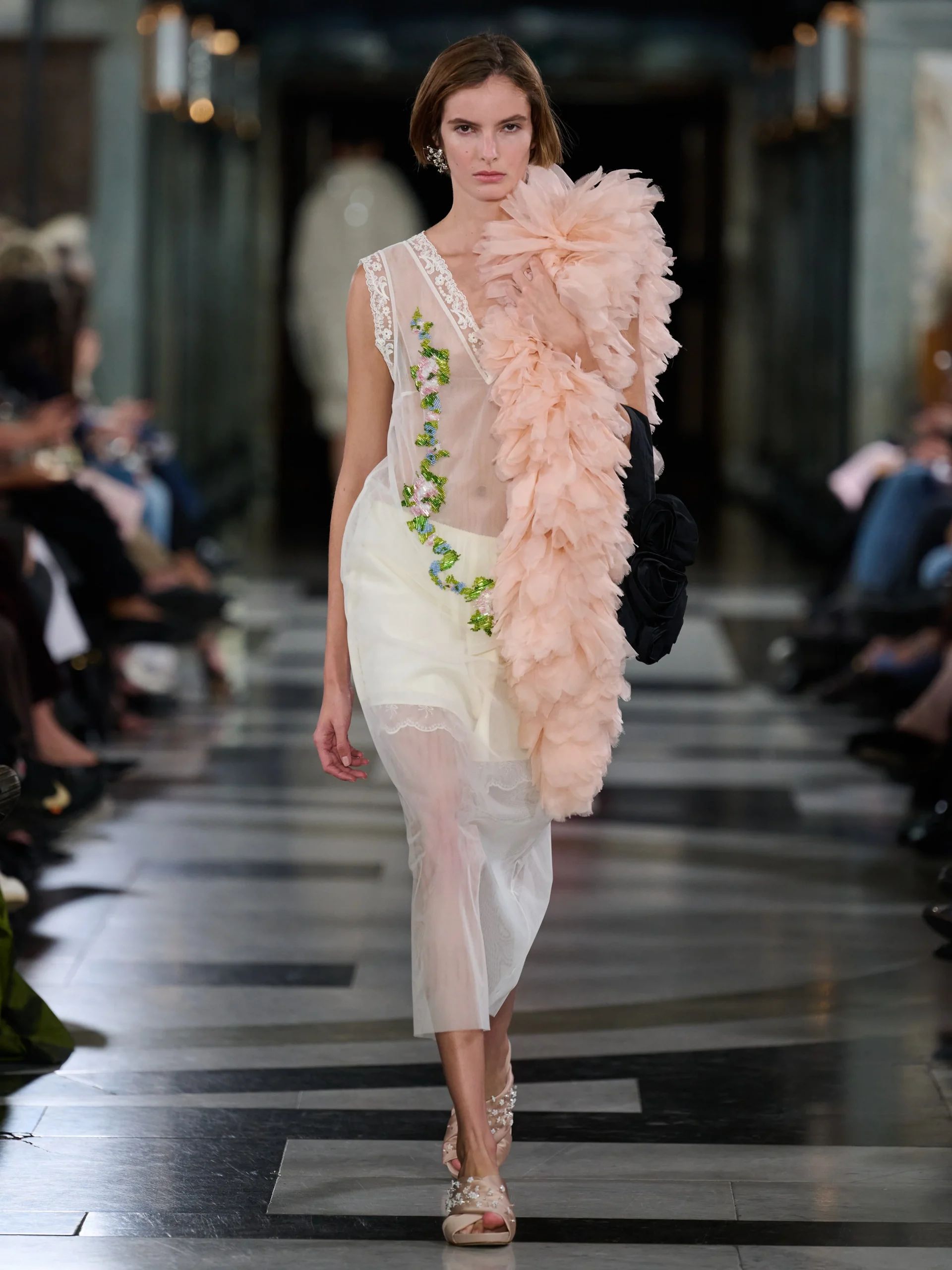 A model walks a catwalk at London Fashion Week, wearing a sheer white dress with green floral accents and a large, fluffy, pale pink frill over one shoulder in a grand hall—an ensemble sure to catch the discerning eye of any fashion editor.