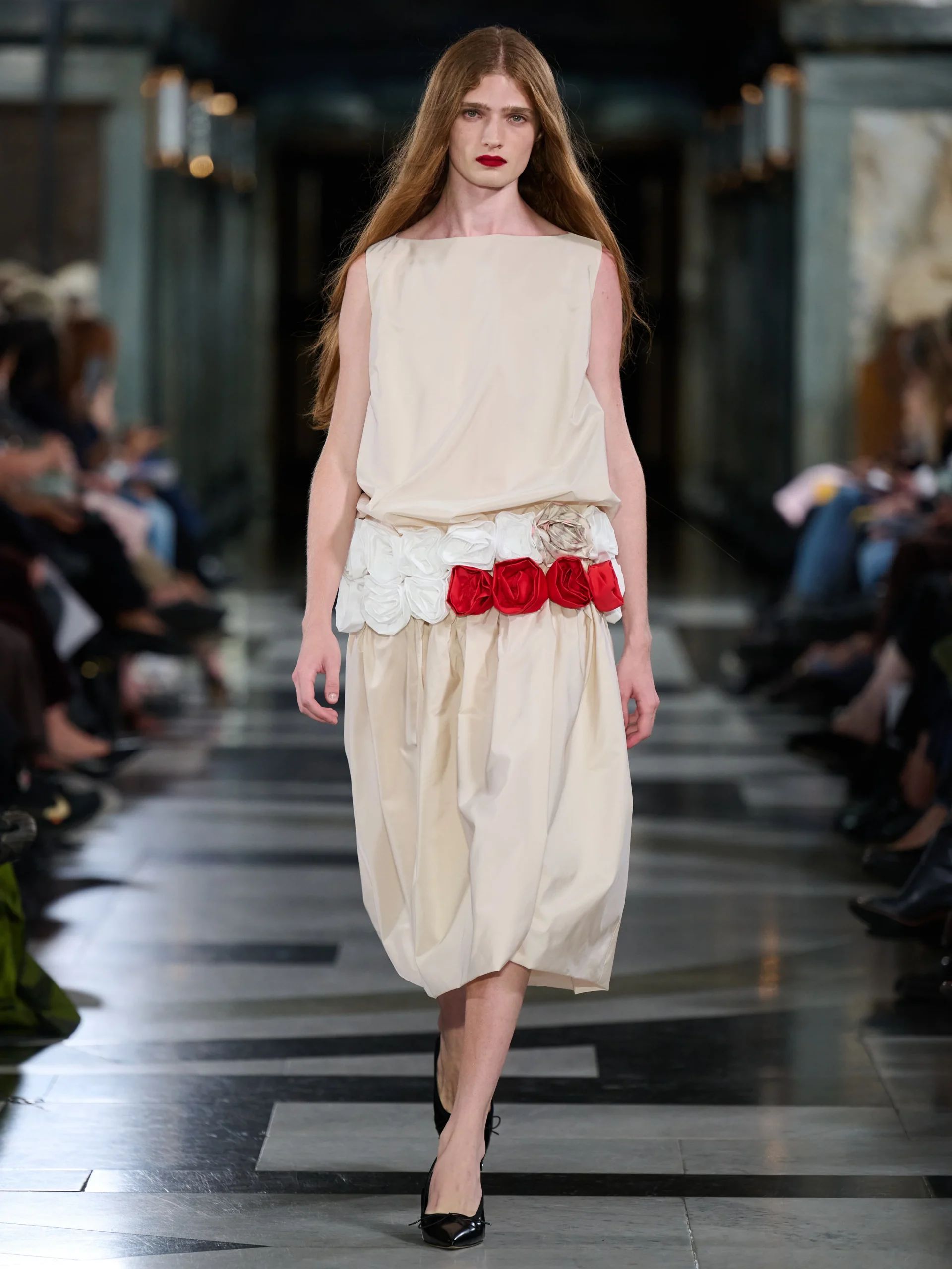 A model walks on a catwalk at London Fashion Week, wearing a cream sleeveless dress with a loose skirt and red and white flower details at the waist, paired with black heels. The background features seated audience and elegant columns.