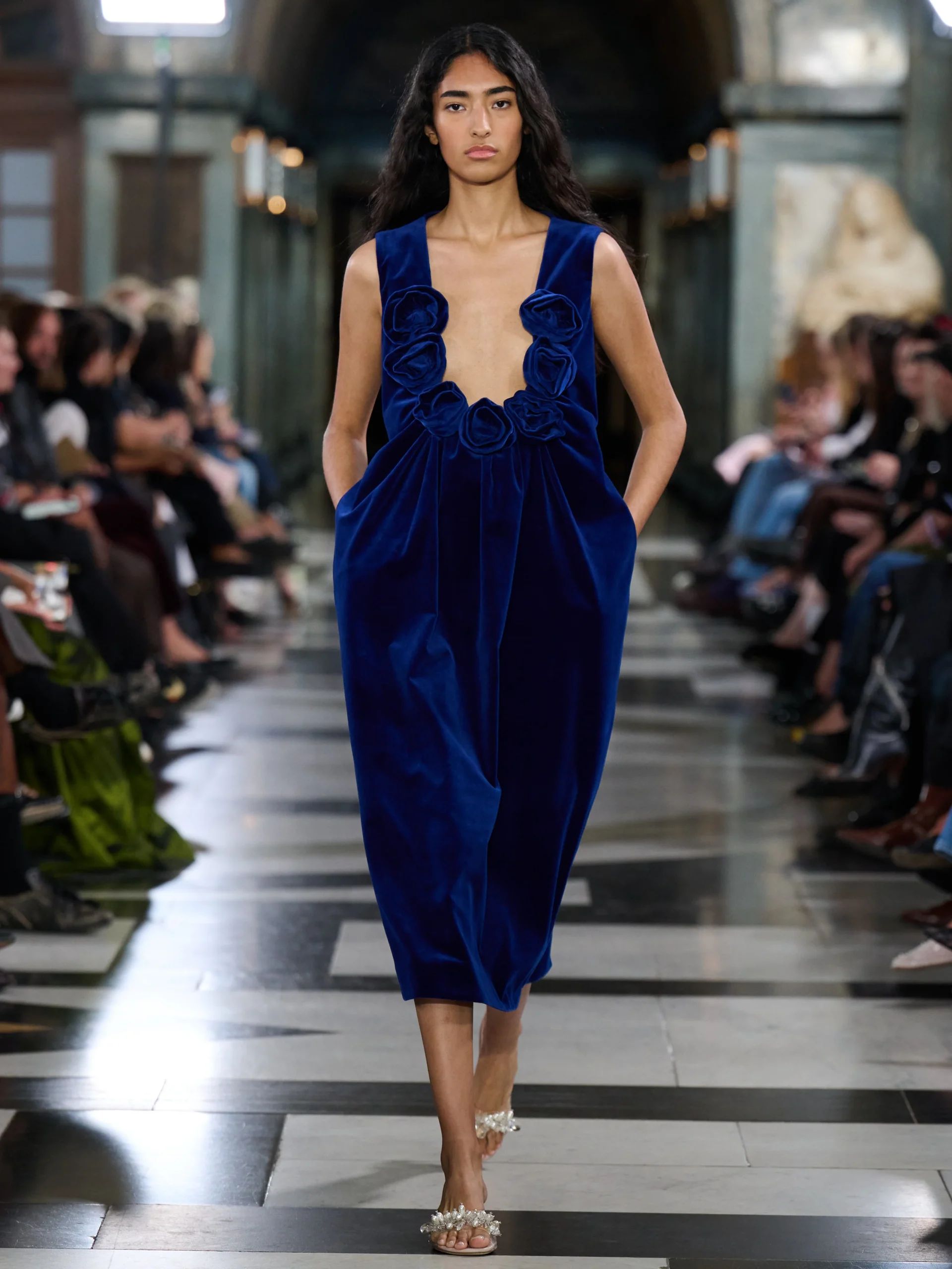 A model walks down a catwalk at London Fashion Week wearing a sleeveless, deep blue velvet dress with decorative frills on the bodice, her hands in the pockets. The audience, including discerning fashion editors, are seated on both sides in a grand hall.