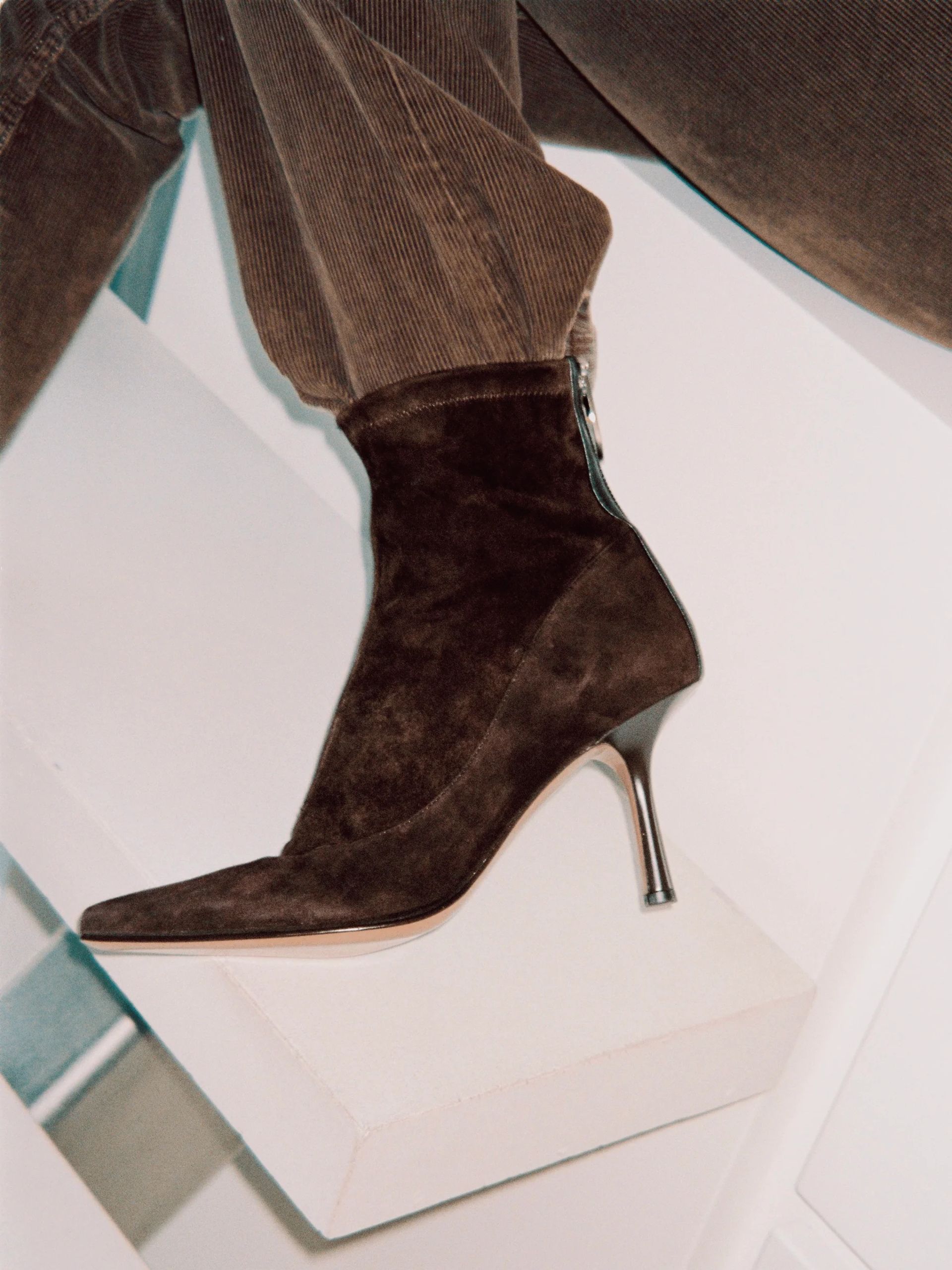 A close-up of a person wearing brown corduroy trousers and a dark brown suede heeled boot with a silver zip and thin heel, foot resting on a white surface—an understated yet sophisticated look often seen from small fashion brands.