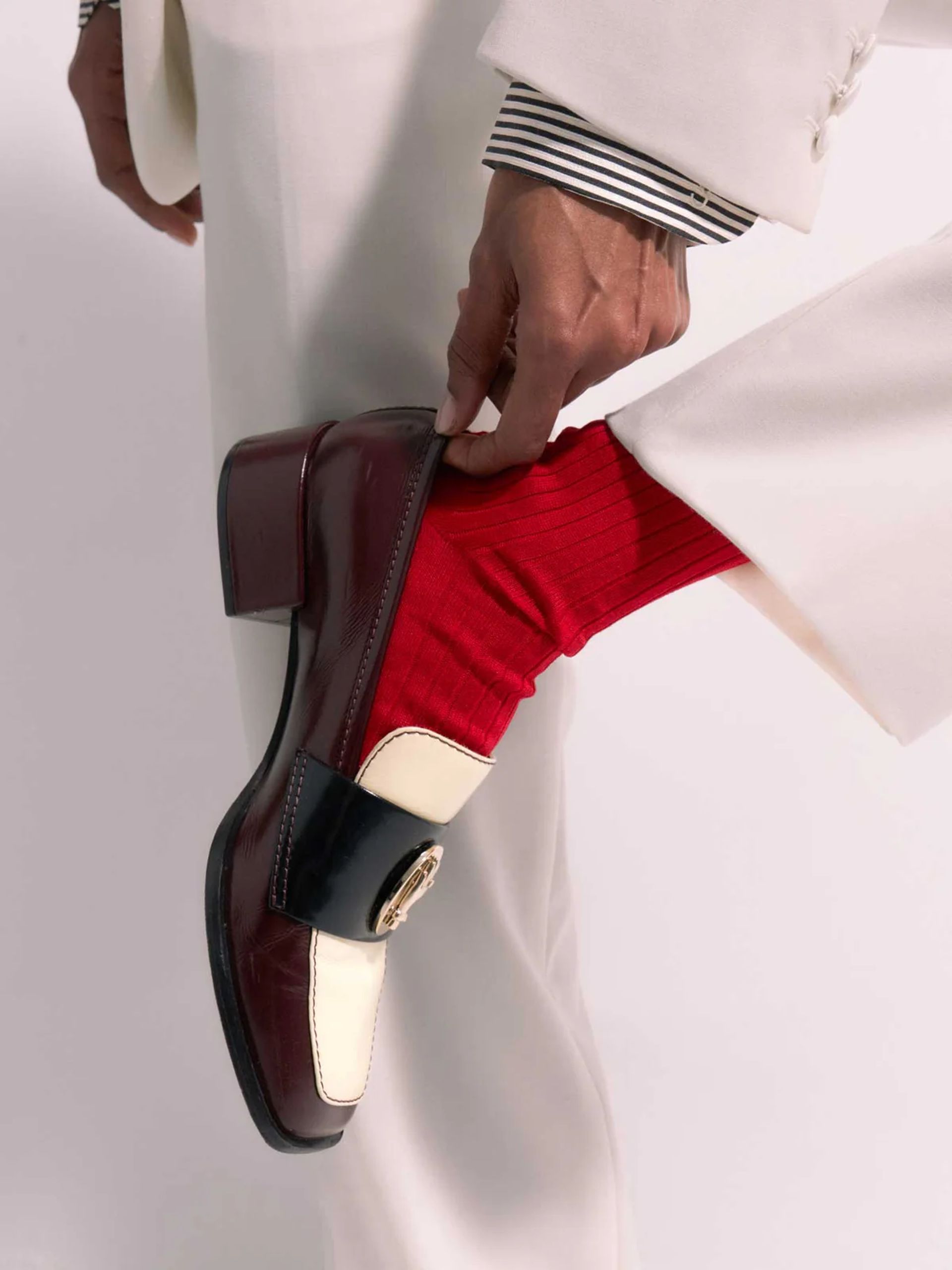A person in a white suit adjusts a red sock, wearing one of the best women’s loafers in burgundy and white with a gold emblem. The outfit includes a striped shirt cuff under the suit sleeve.