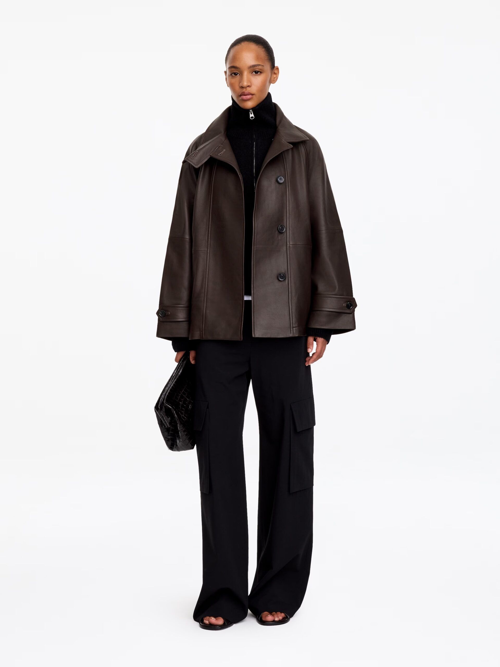 A model wearing one of the best women's leather jackets, styled with a black polo neck, black cargo trousers, and a black clutch.
