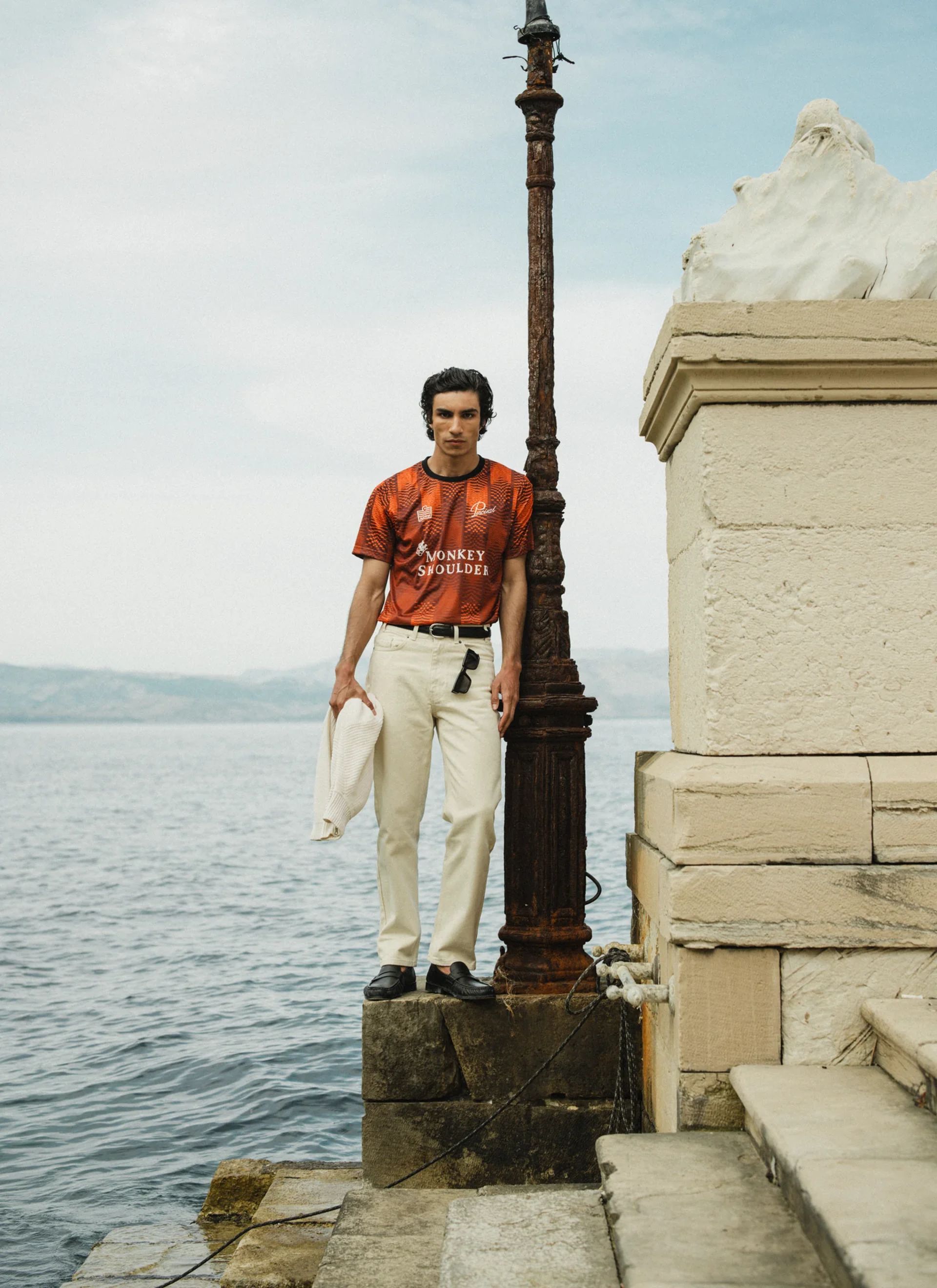A man in an orange football core graphic t-shirt and white trousers stands on a stone platform by the water, holding a white jacket. An old lamp post and stone structure are beside him, with the sea and distant hills in the background.