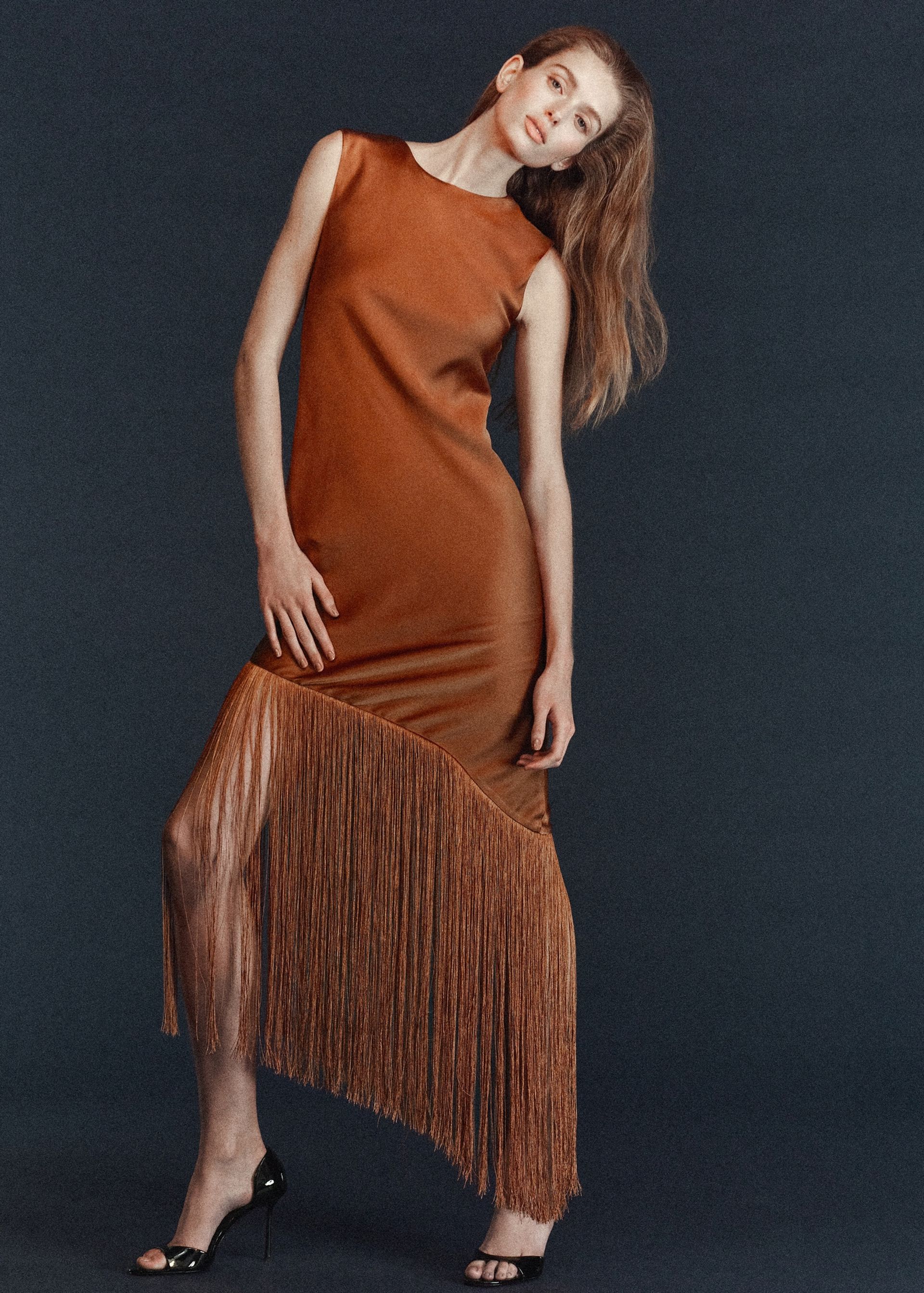 A woman poses in a sleeveless, rust-coloured dress with an asymmetrical fringed hem, showcasing her stylish fringe benefits against a dark background whilst wearing black high-heeled sandals.