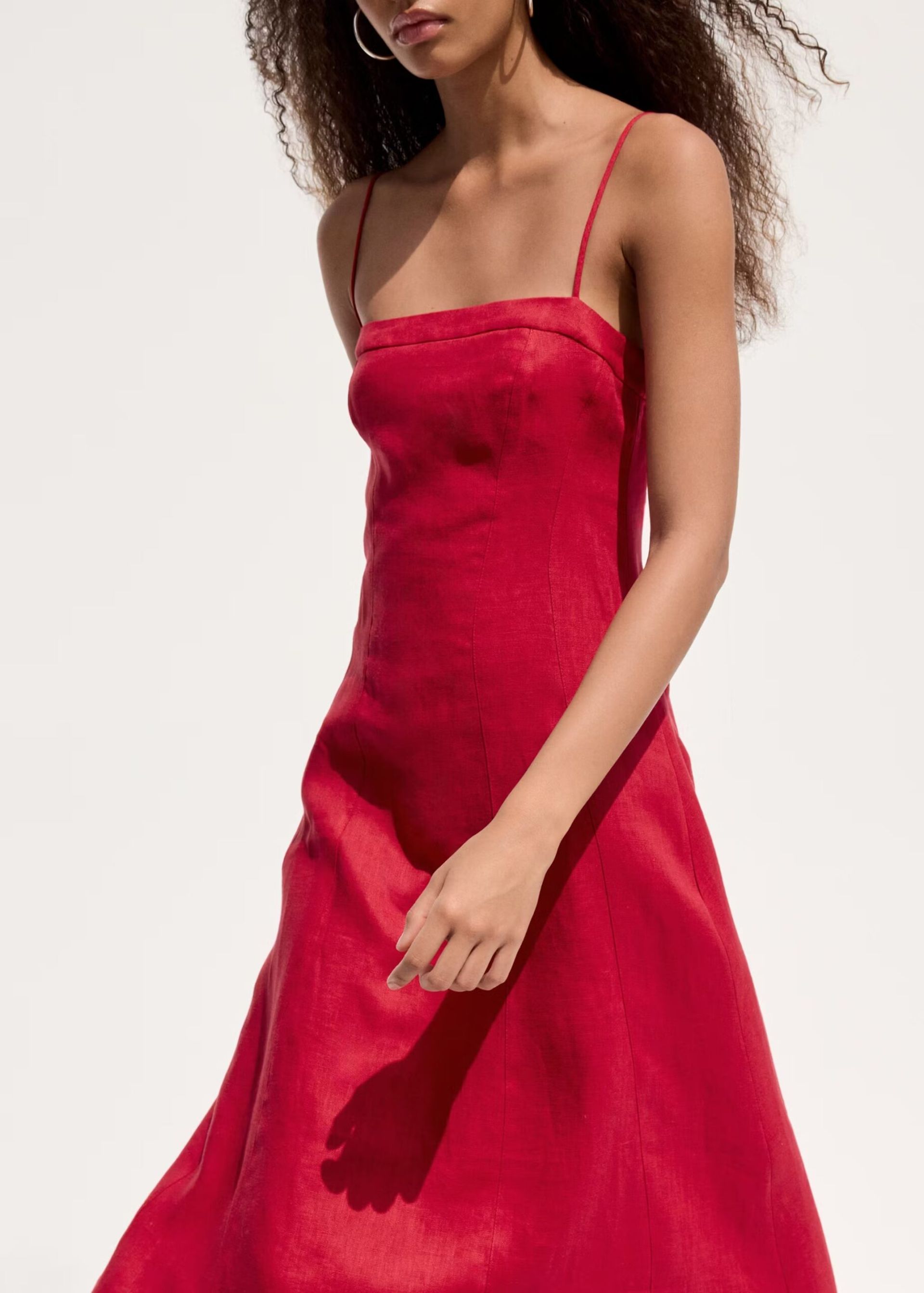 A woman with curly hair wears a sleeveless, bright red high street linen dress with thin straps and a straight neckline, walking against a plain, light background.