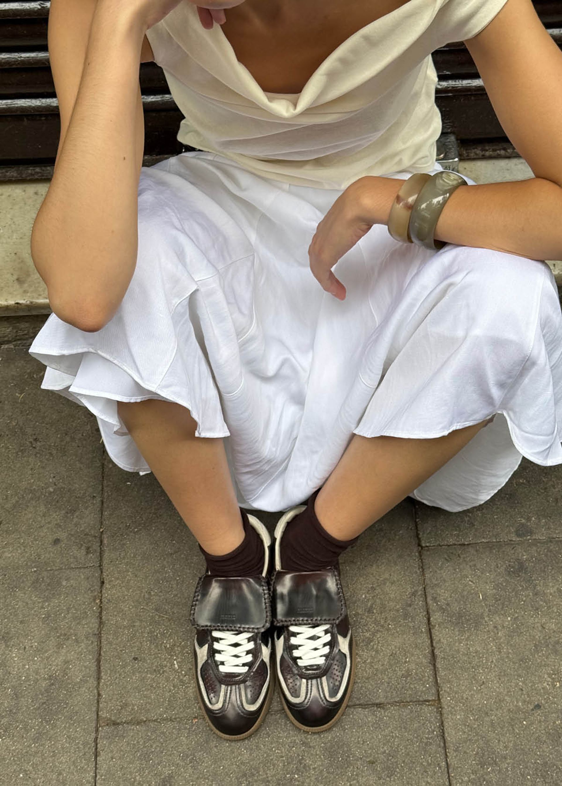 A person in a white dress sits on a pavement, wearing retro football style trainers, brown socks, and chunky bracelets, with one arm resting on their knee.