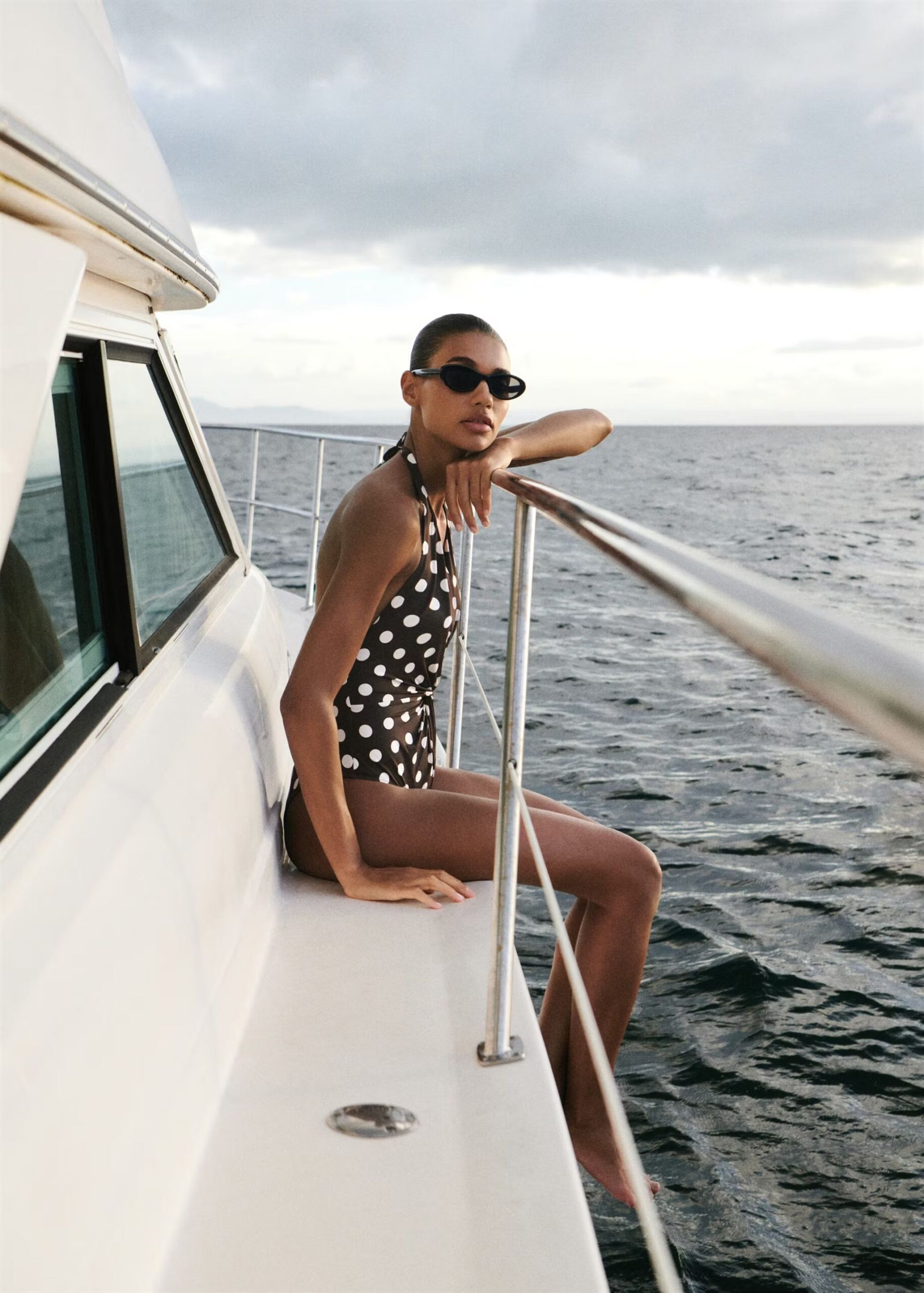 A woman in pretty swimwear—a black-and-white polka dot swimming costume—sits on the edge of a boat, wearing sunglasses, with calm sea water and a cloudy sky in the background.