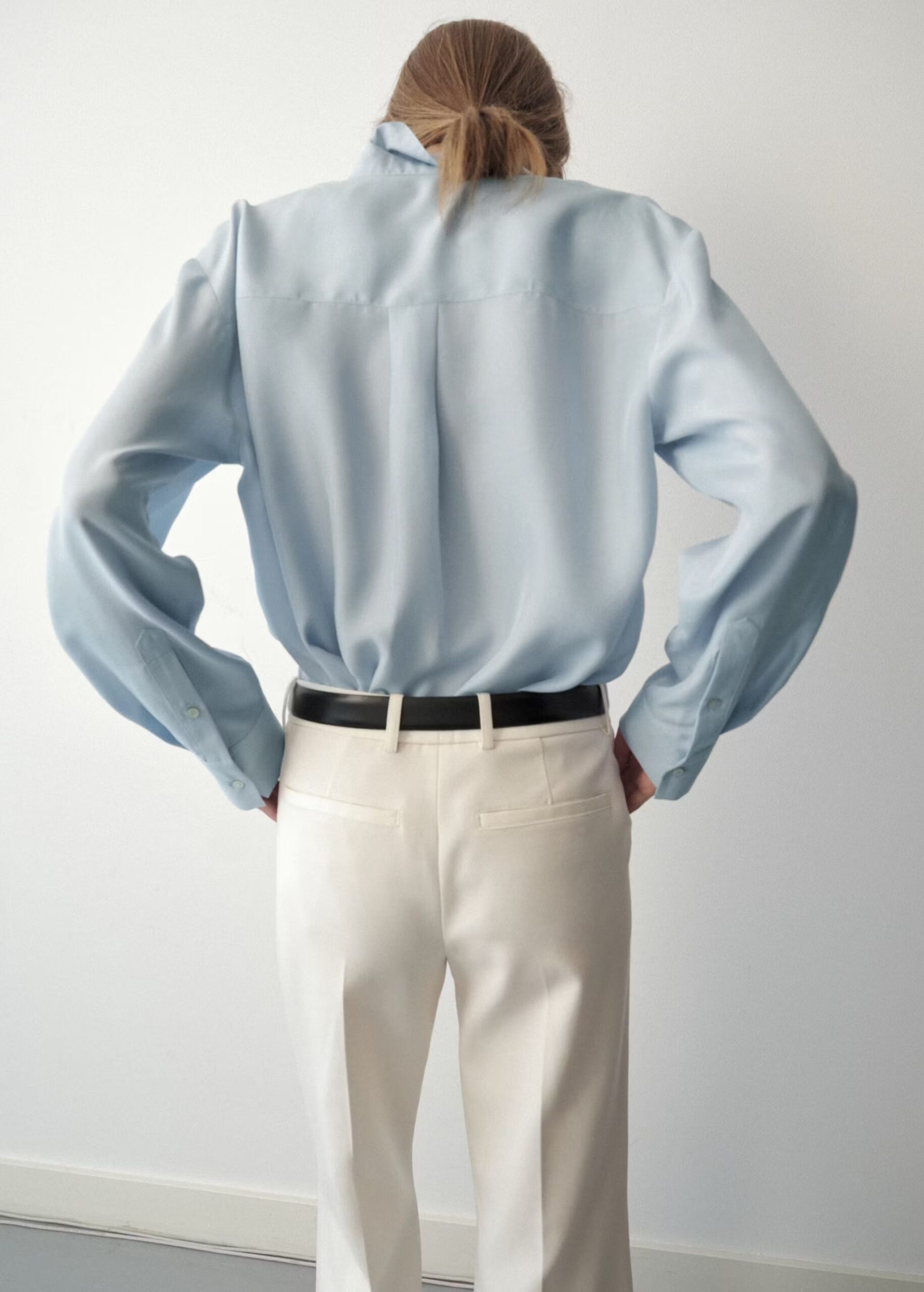 A person with light brown hair in a ponytail stands facing away, wearing a light blue button-up shirt tucked into high-waisted white trousers with a black belt—a look reminiscent of minimalist high street brands. The background is a plain, light-coloured wall.