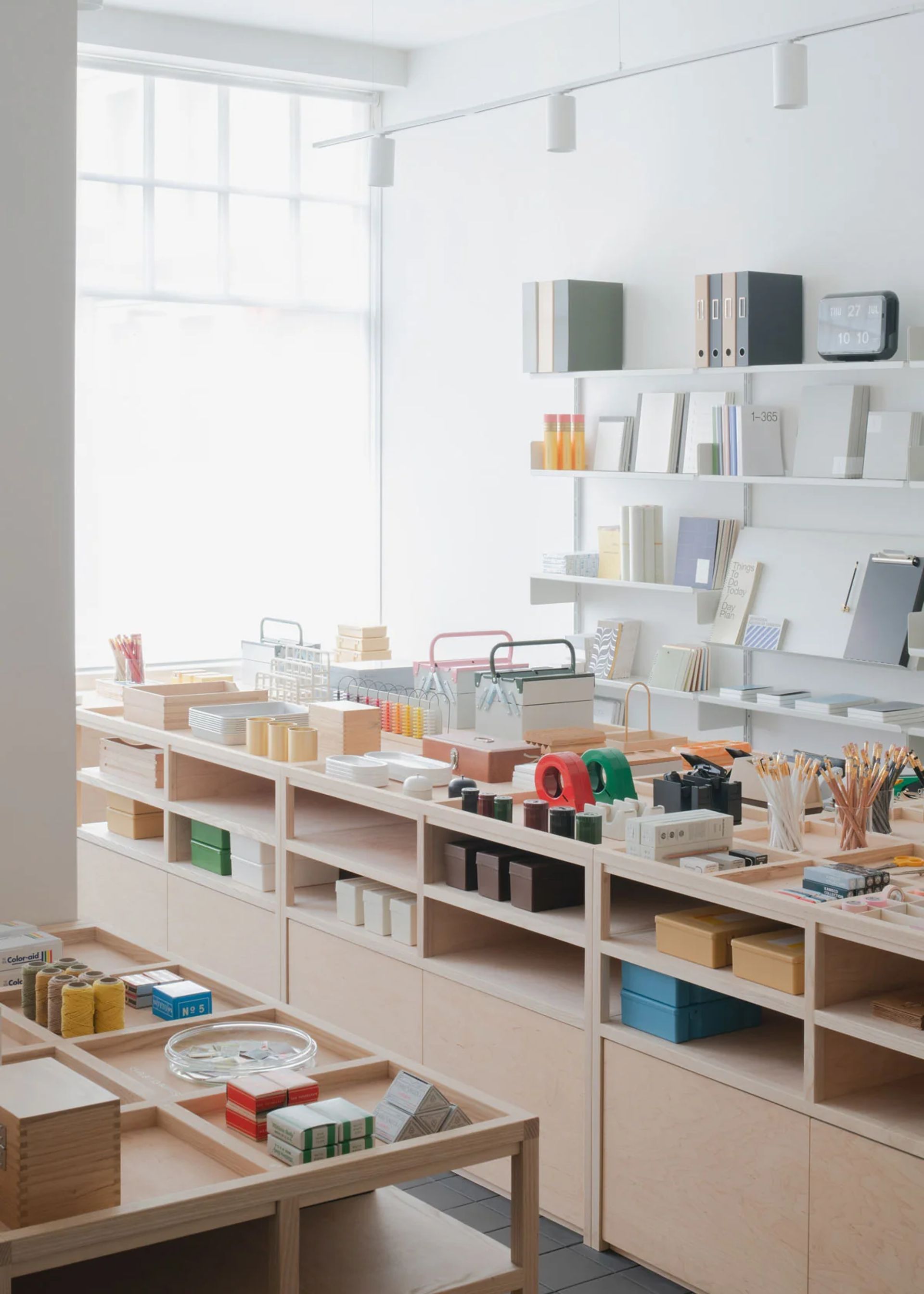 A bright, minimalist stationery shop with organised wooden shelves displays notebooks, tapes, pens, and other office supplies in natural light from a large window—a top pick among local stationery shops.
