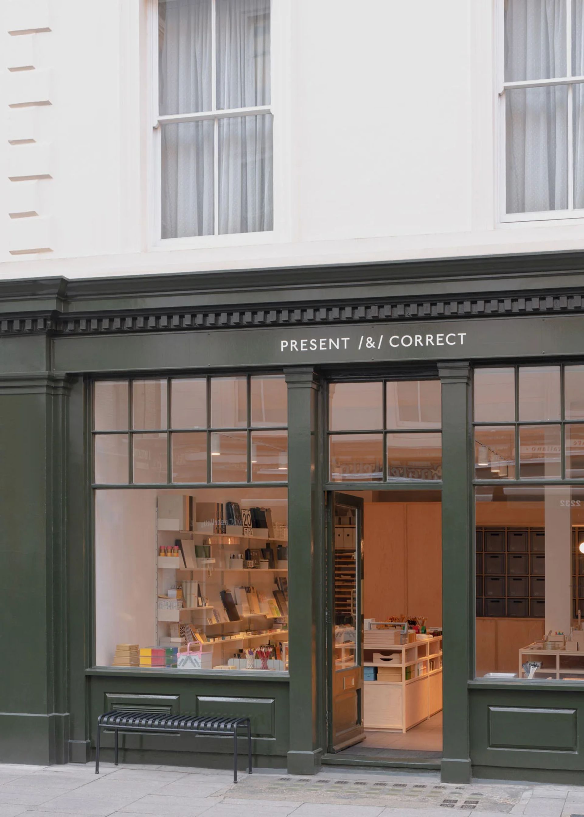A stationery shop with dark green trim and large windows displays neatly arranged office supplies inside. The shop sign above the door reads PRESENT & CORRECT. A black bench sits on the pavement outside.