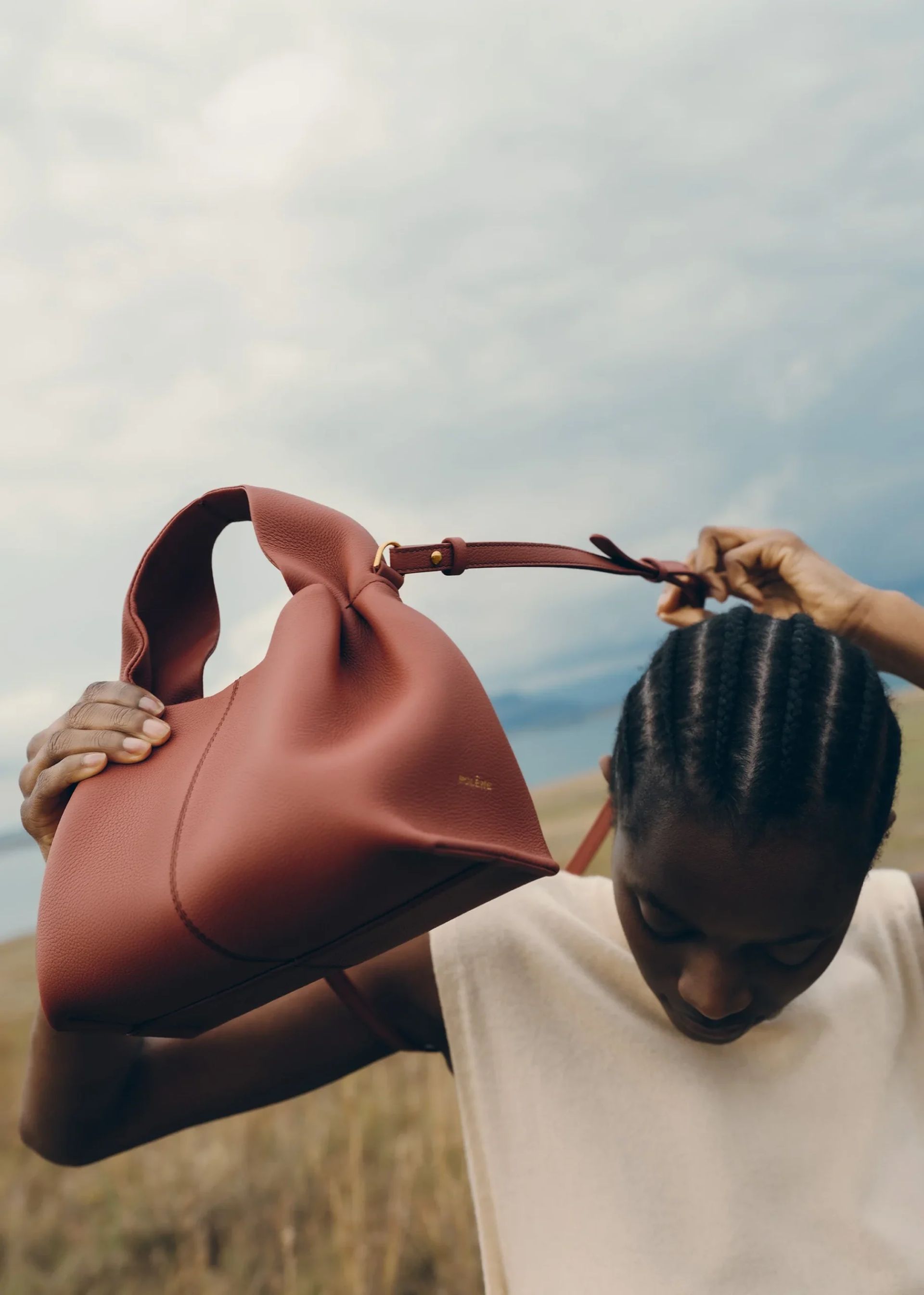 A person with plaited hair holds a large, soft, brown designer bag above their head outdoors, looking down. The background is blurred, with sky and landscape visible.