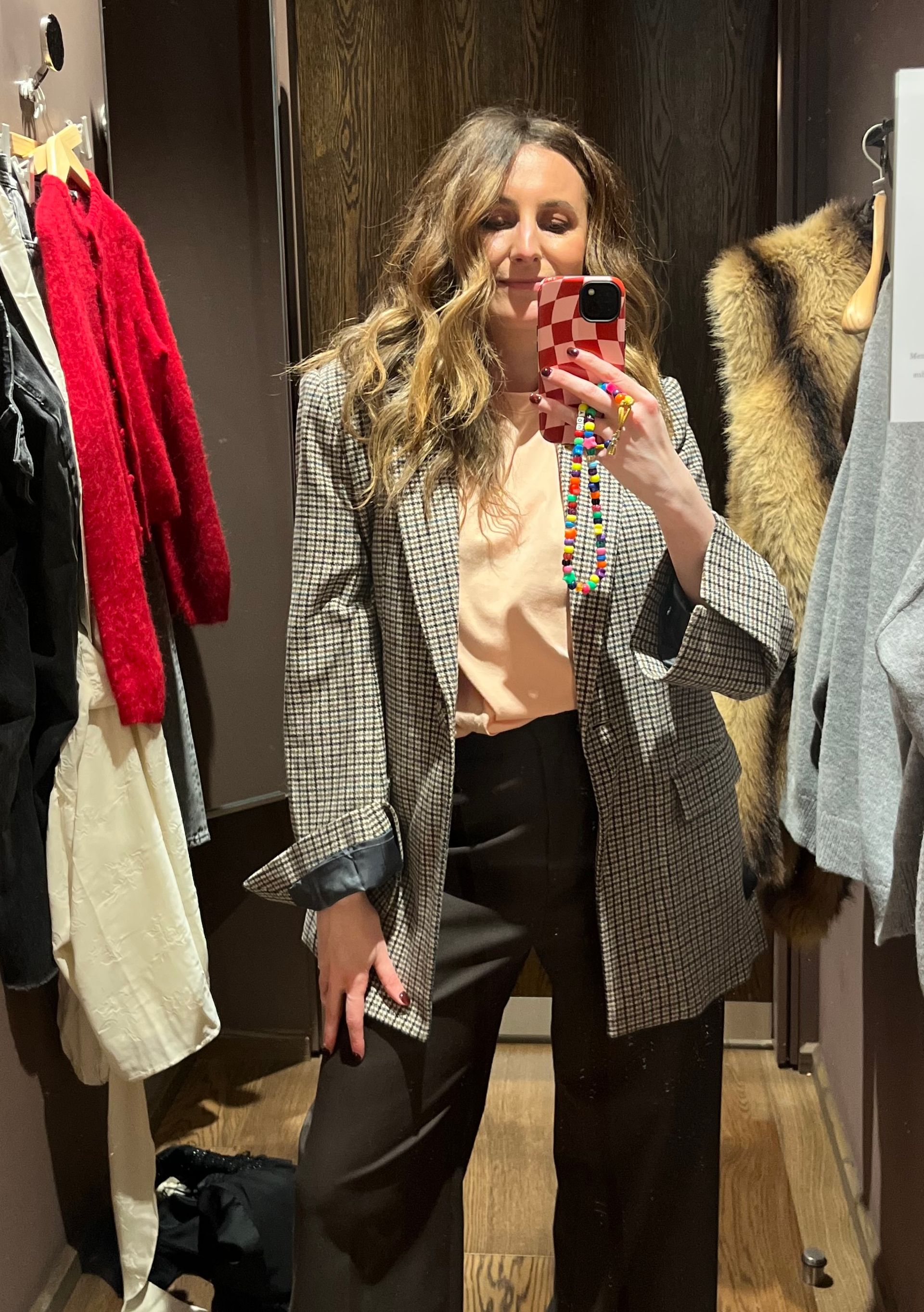 A woman with wavy hair takes a mirror selfie in a changing room, wearing a High Street Blazers checked jacket over a peach top and dark trousers. Various clothes hang on hooks, and a faux fur coat adds Fashion Editor flair to the background.