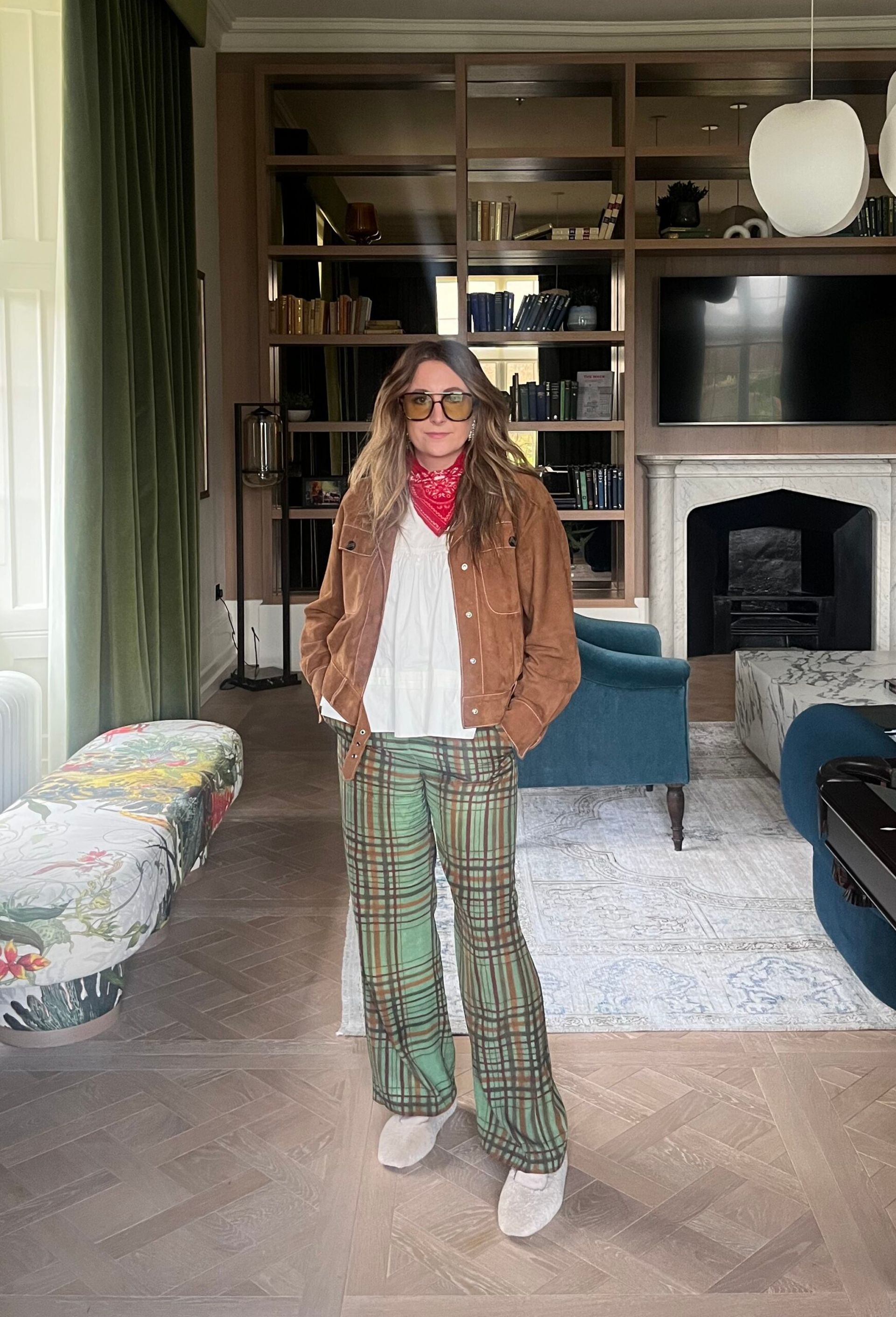 A woman stands in a stylish living room with bookshelves and a fireplace, wearing a chic brown suede jacket and green checked trousers—recreate outfits like this or shop my wardrobe for similar pieces.