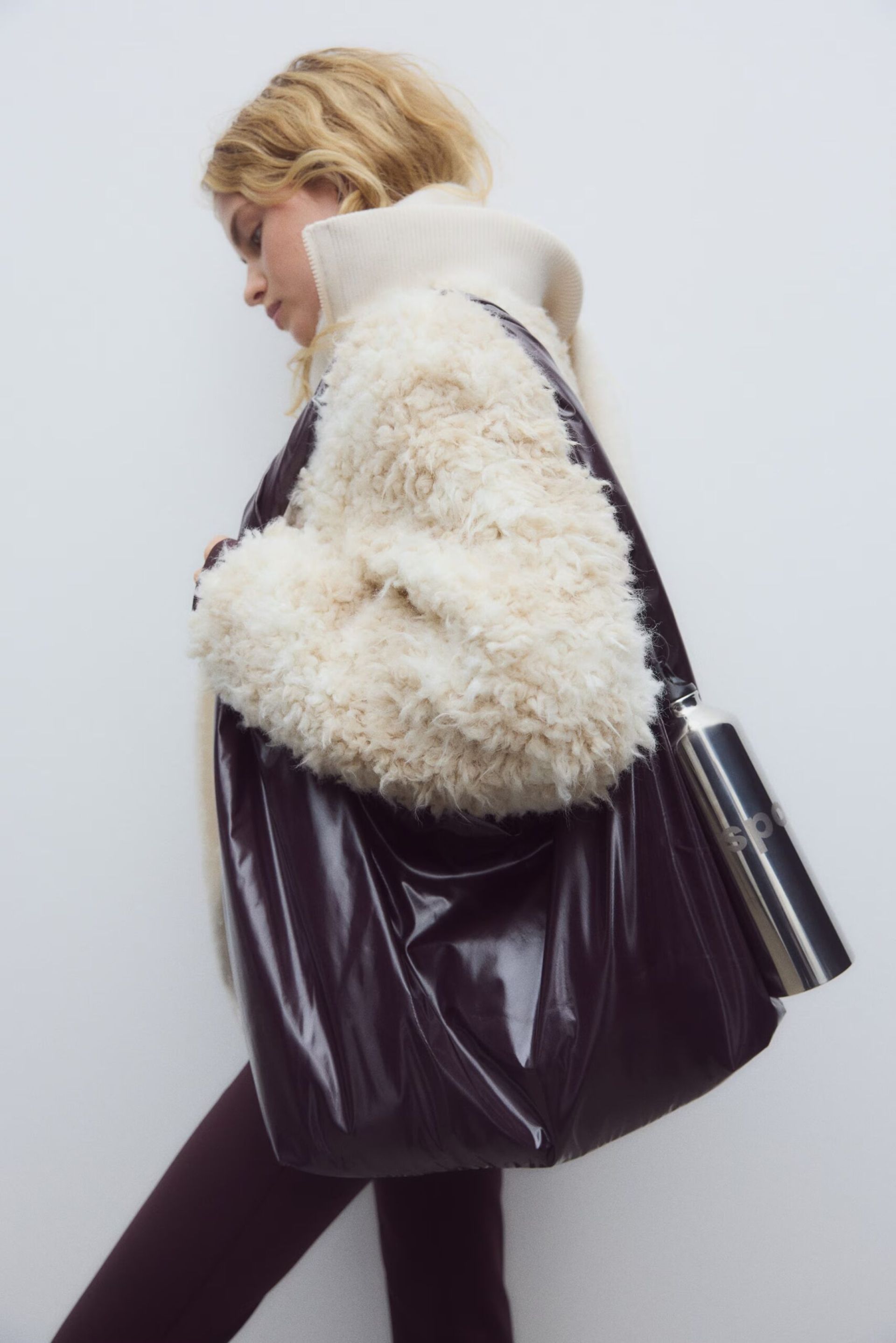 A person with blonde hair wears a cream, fluffy jacket styled with the latest sportswear and carries a large, shiny dark brown tote bag with a metal water bottle in the side pocket, standing against a plain white background.