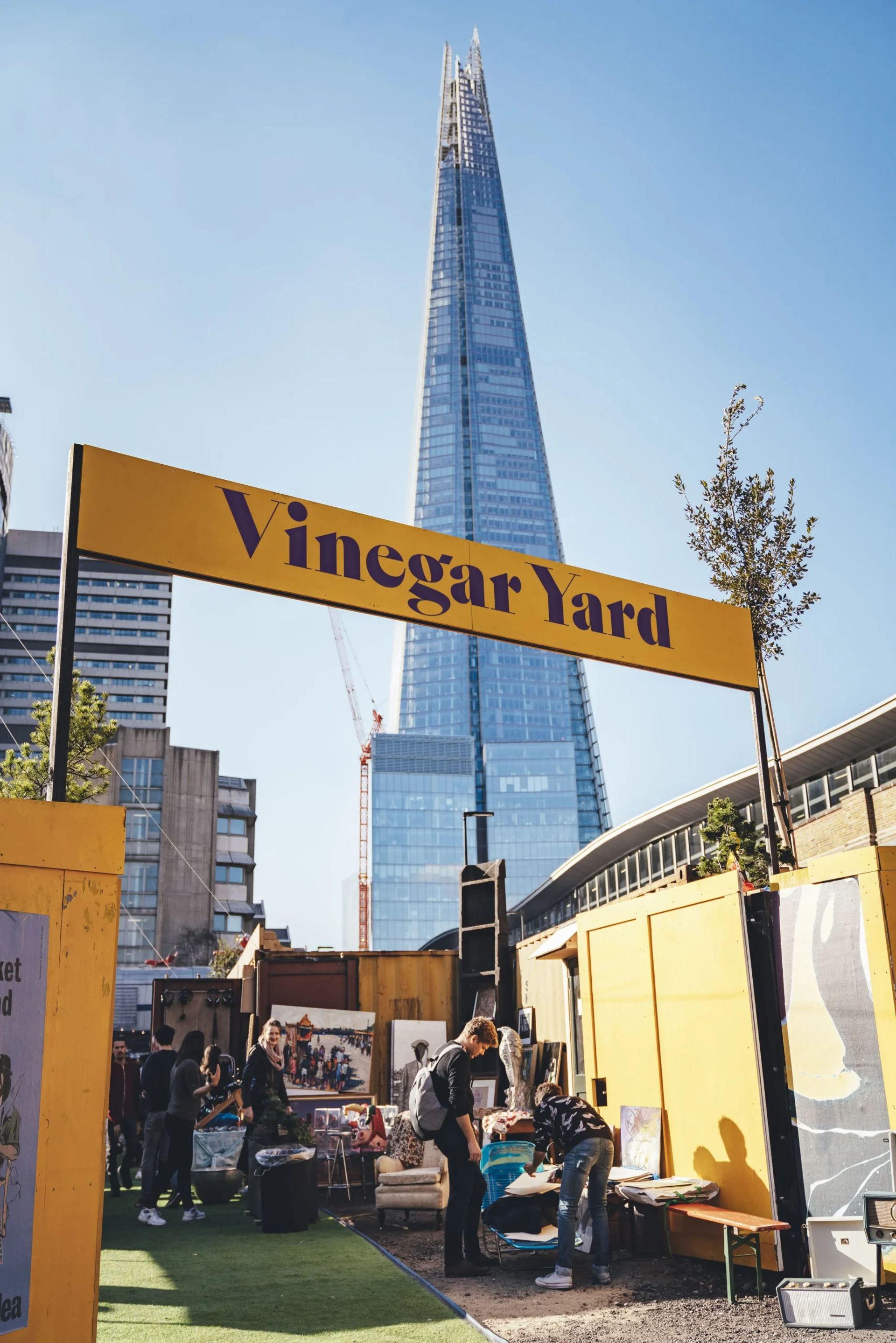 A yellow sign reads Vinegar Yard at an outdoor market—a must-visit in any London flea market guide—with people browsing stalls. The Shard skyscraper rises in the background under a clear blue sky.