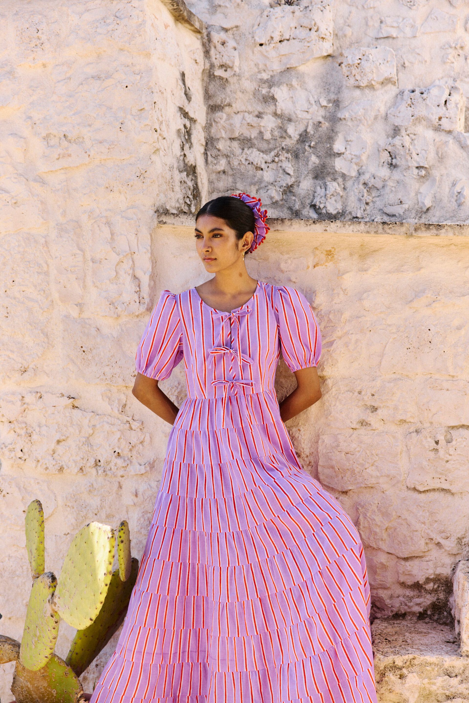 A woman in a long, pink and red striped dress stands against a textured stone wall, with a cactus in the foreground. Her stylish look is perfect inspiration for your weekend activities or to feature in your next weekend guide.