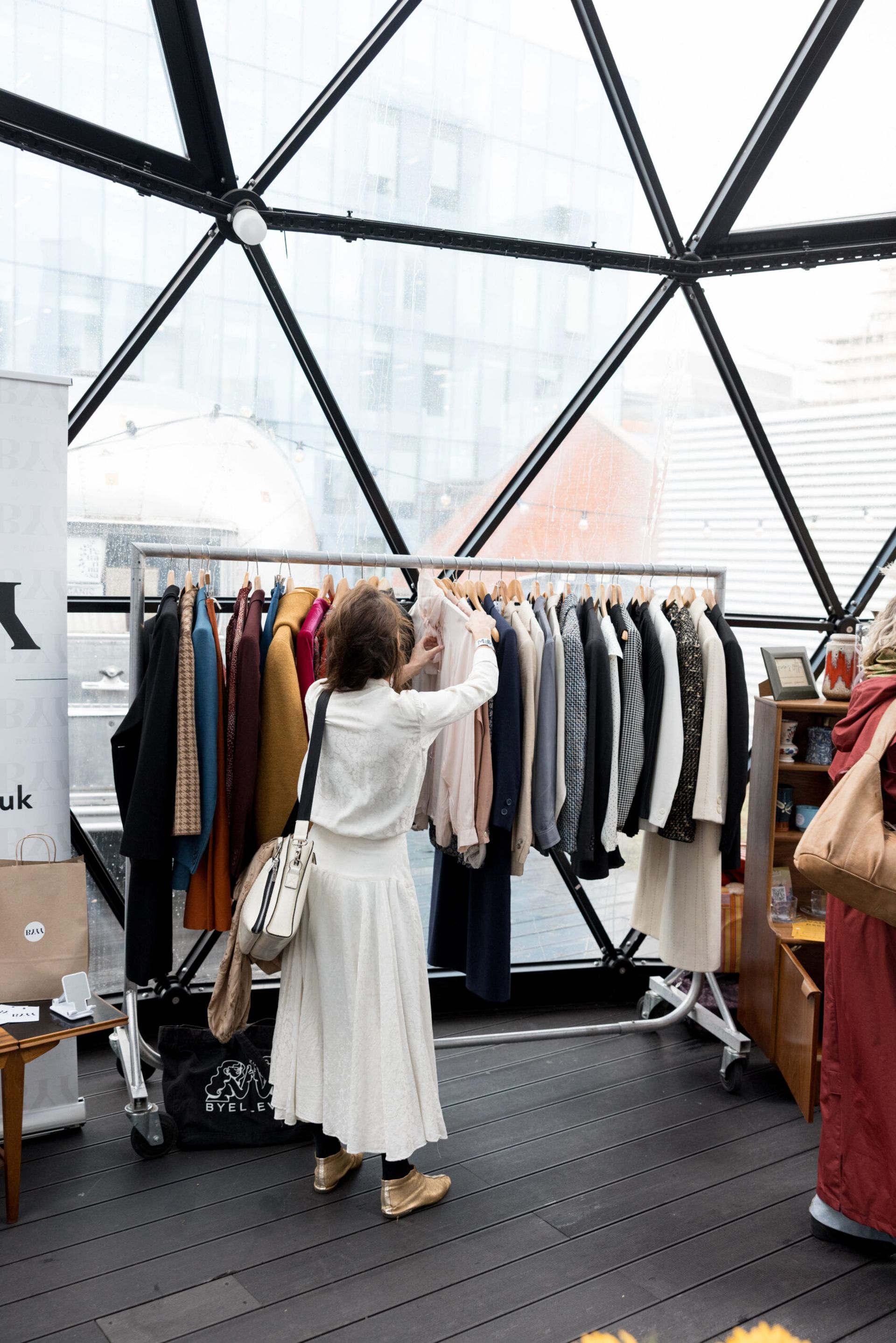 A woman in a white outfit browses a rail of assorted coats and jackets inside a modern glass-domed structure, city buildings in the background—a perfect scene for any WEEKEND GUIDE.