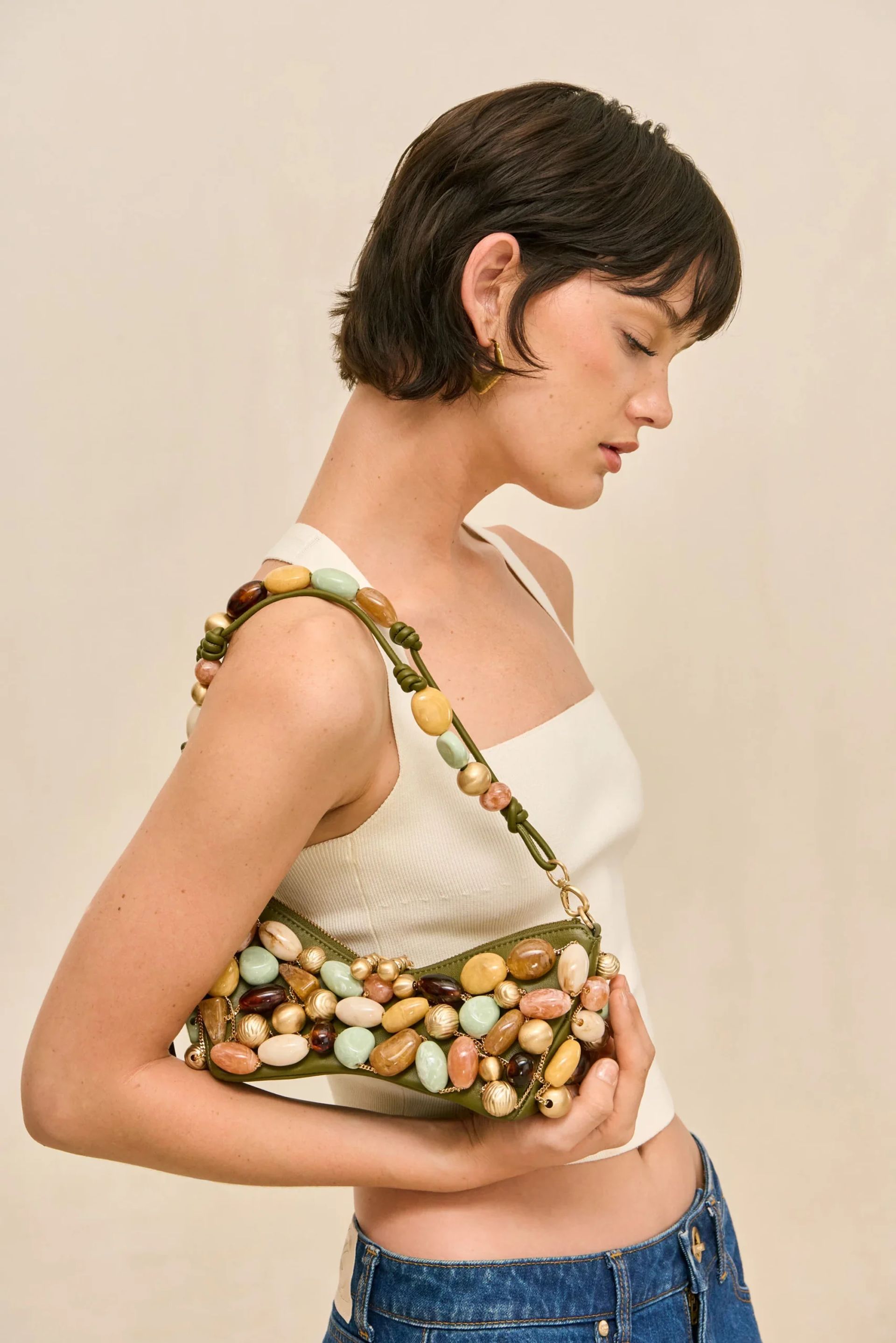 A woman with short dark hair, in a white sleeveless top and blue jeans, holds a stylish beaded bag over her shoulder, its large colourful beads standing out against the plain beige background.