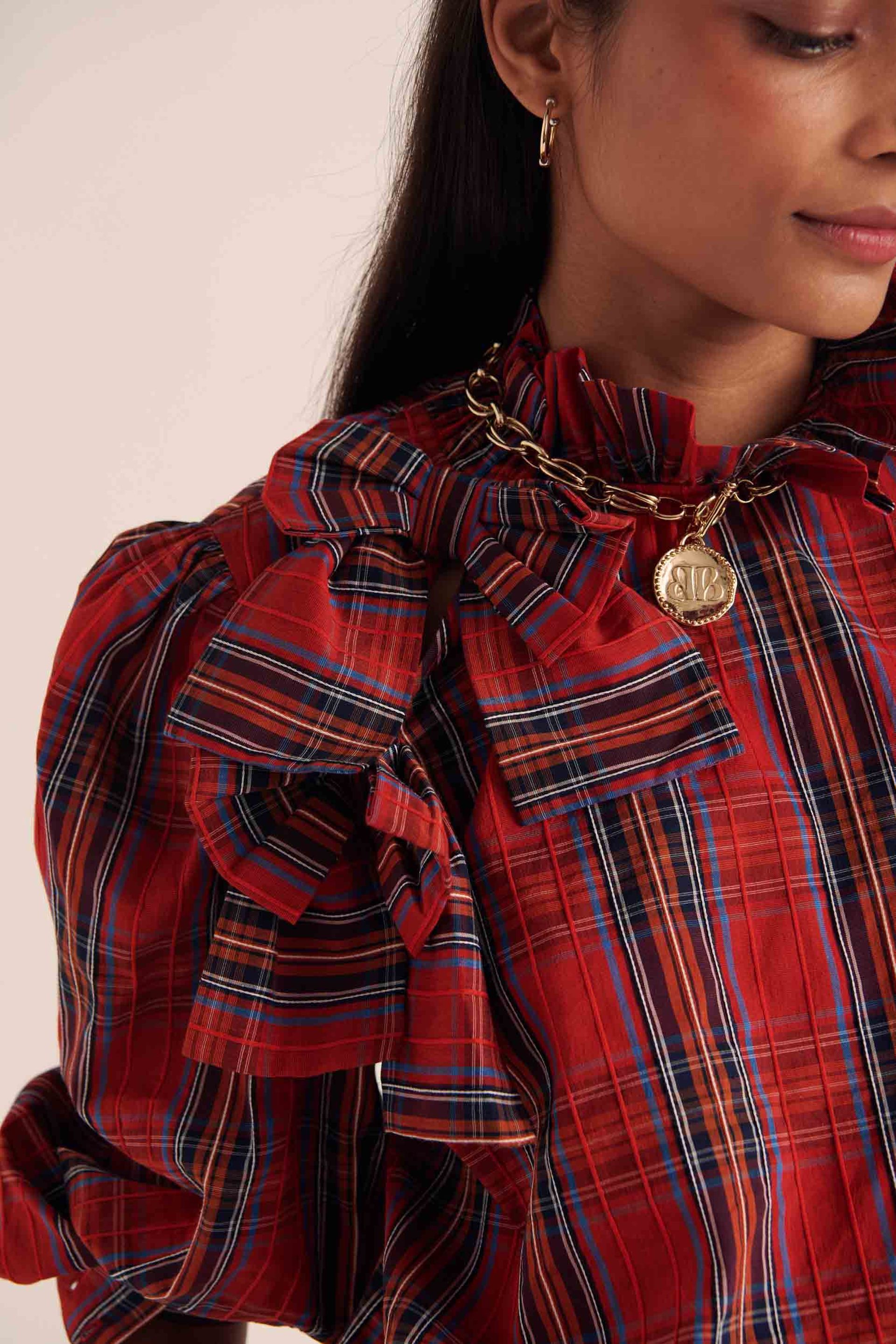 Close-up of a woman wearing a red checked blouse with large bows on the shoulders, embracing bow fashion. She accessorises with a gold chain necklace featuring a round pendant and a small hoop earring. Her face is partially visible, showing a slight smile.