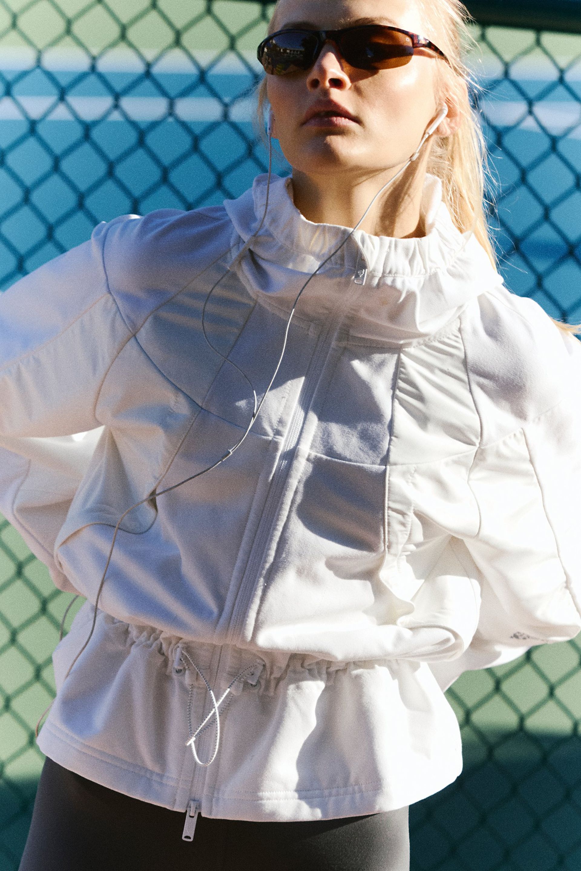 A person wearing the latest sportswear—a white zip-up jacket, sunglasses, and earphones—stands outside in sunlight in front of a green chain-link fence.