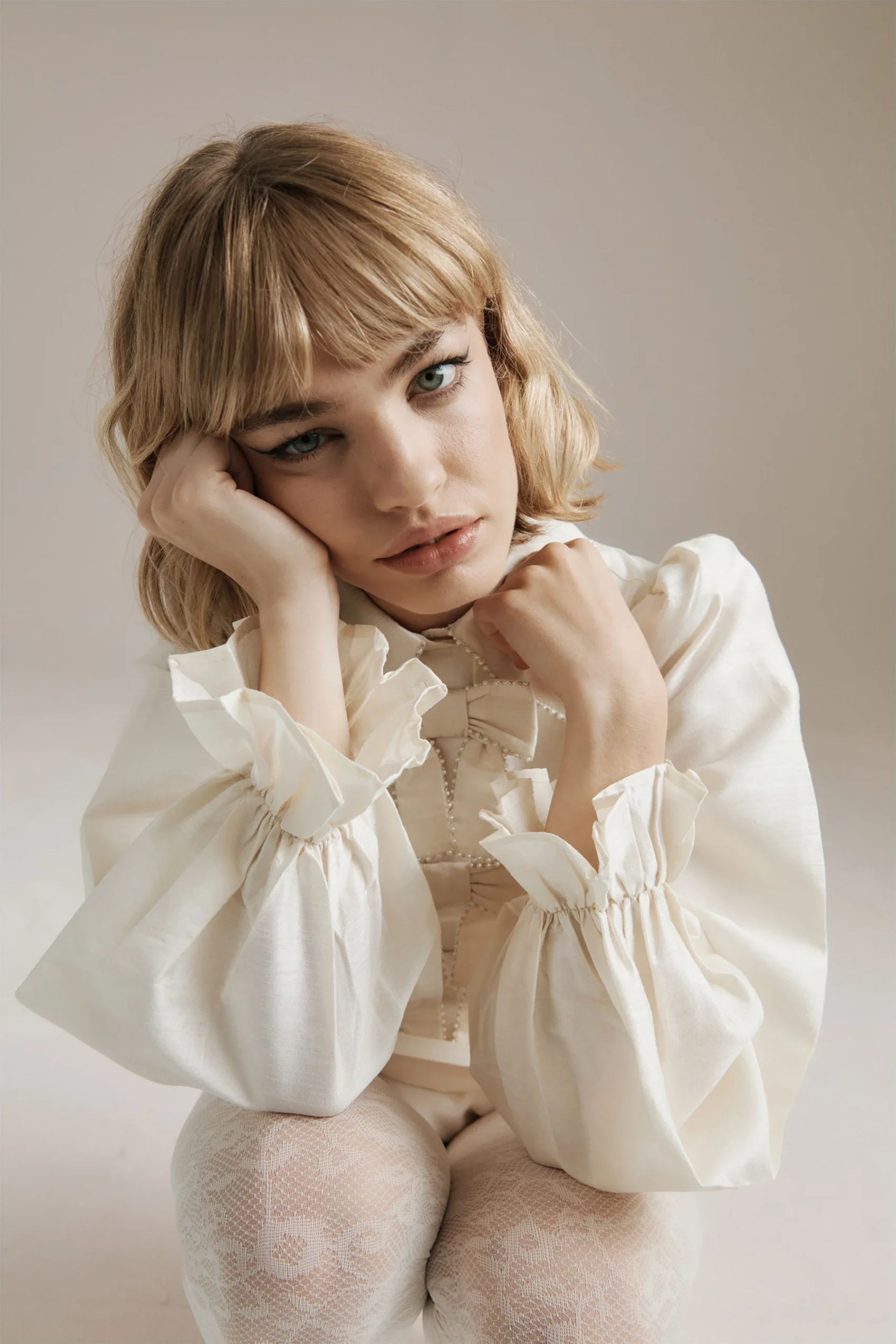A young woman with blonde hair and fringe, wearing a white blouse with frilled sleeves and a subtle bow fashion accent, rests her head on her hand and looks directly at the camera with a neutral expression against a plain background.