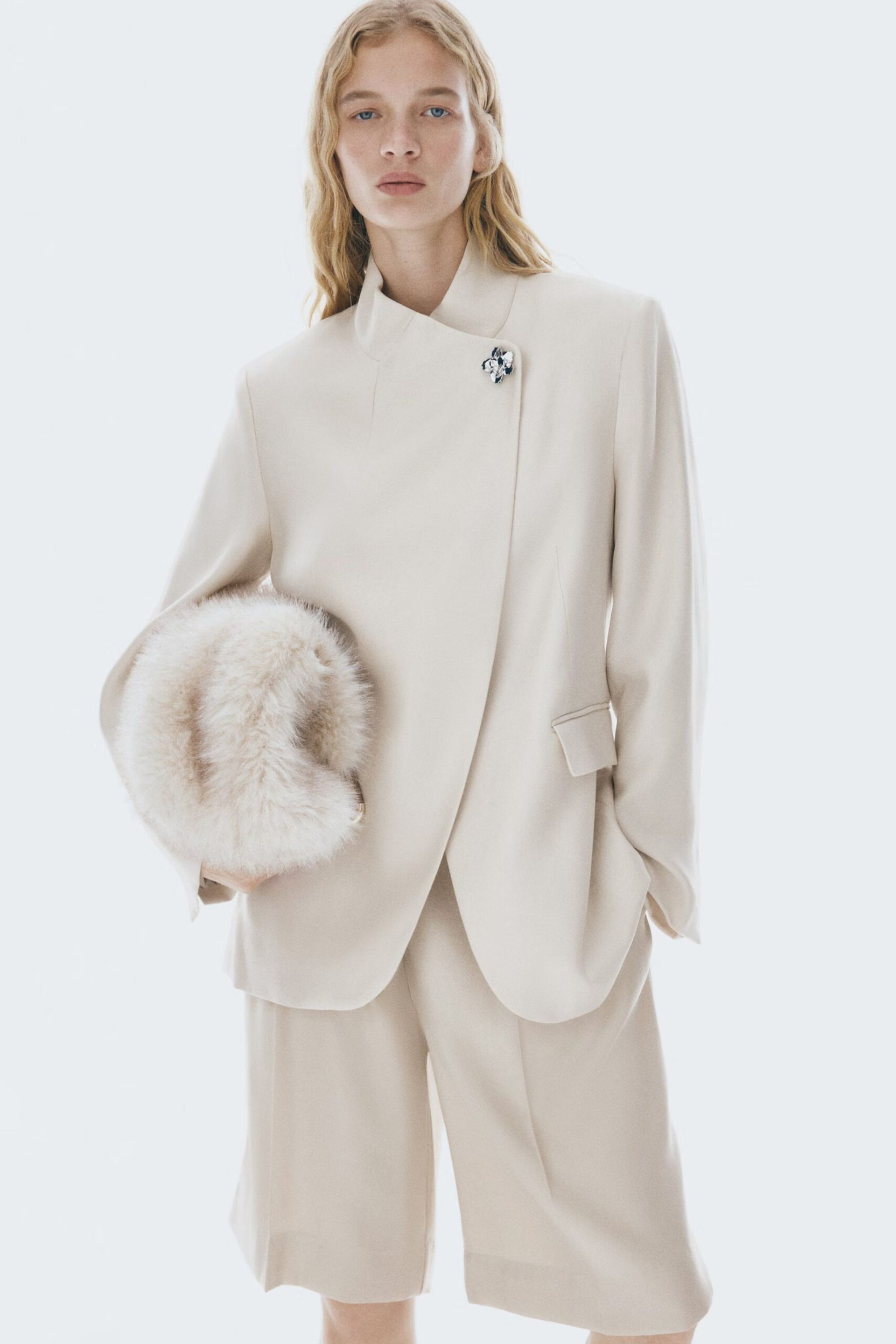 A person with long blonde hair wears a cream-coloured High Street blazer and matching knee-length shorts, holding a fluffy, light accessory. The blazer features an asymmetrical design with a decorative brooch near the collar.
