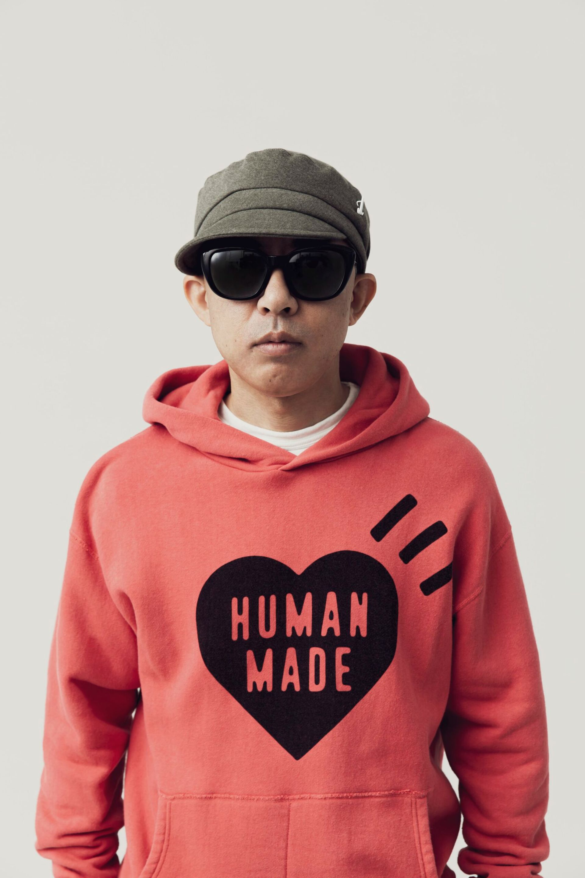 A person wearing a red hoodie with a black heart design and the words HUMAN MADE, dark sunglasses, and a grey cap stands against a white background—ready to turn heads at fashion events.