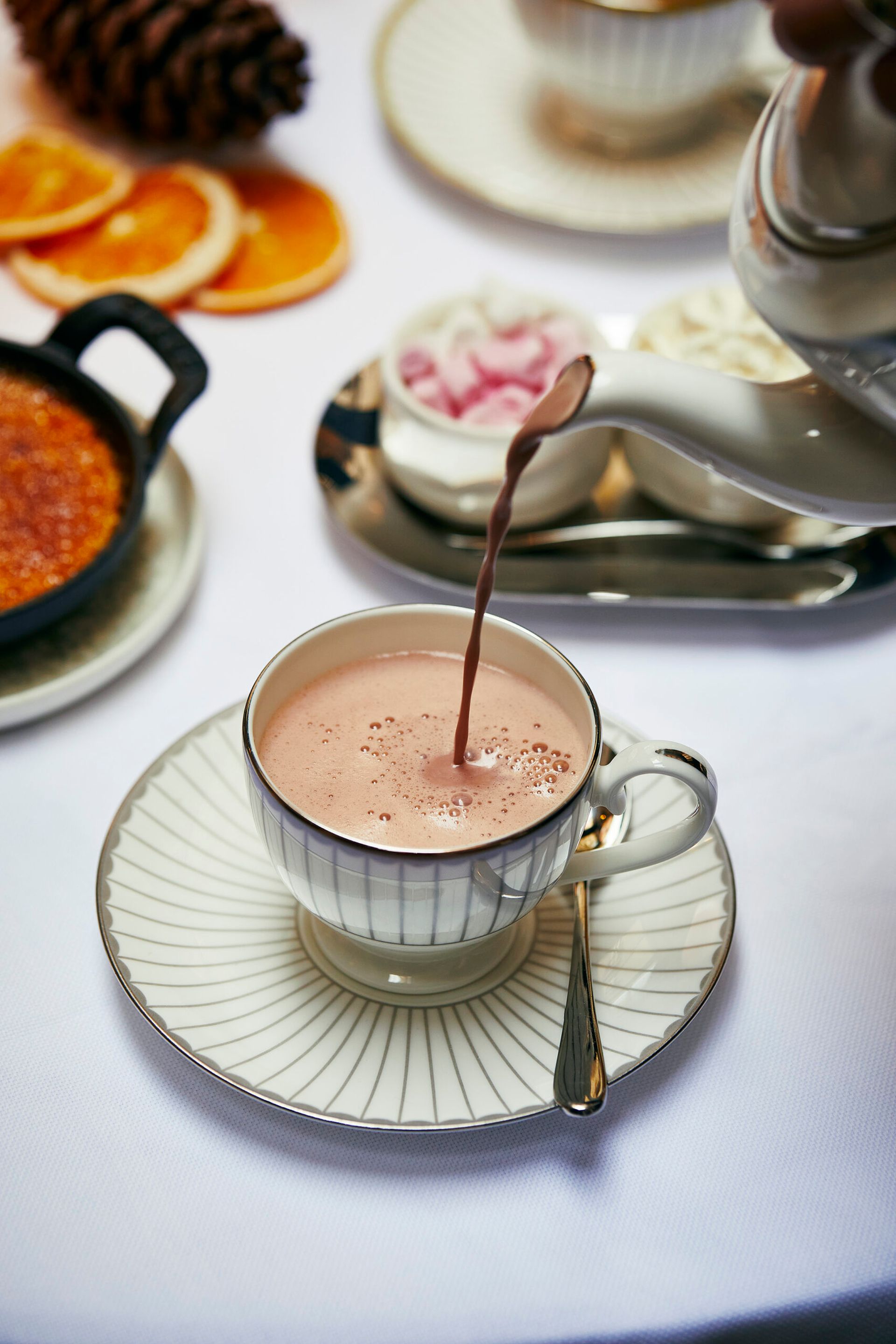 A cup of hot chocolate, one of the classic winter drinks, is poured from a teapot into an elegant teacup with a spoon on the saucer. In the background are sugar lumps, orange slices, and a pudding on a white tablecloth.