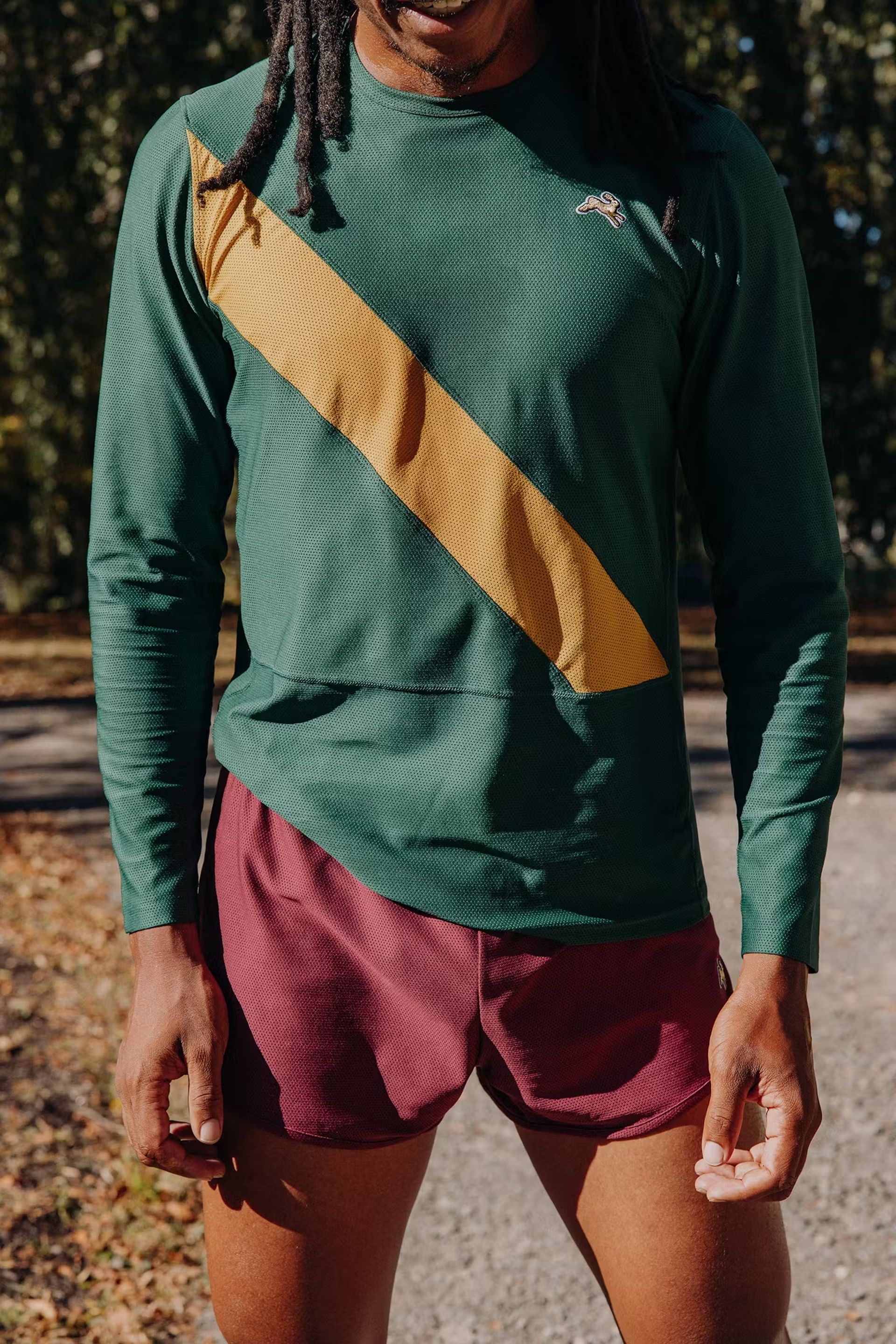 A person wearing men's gym clothes—a green long-sleeved shirt with a diagonal yellow stripe and maroon athletic shorts—stands outdoors on a sunny day, with trees and fallen leaves in the background.