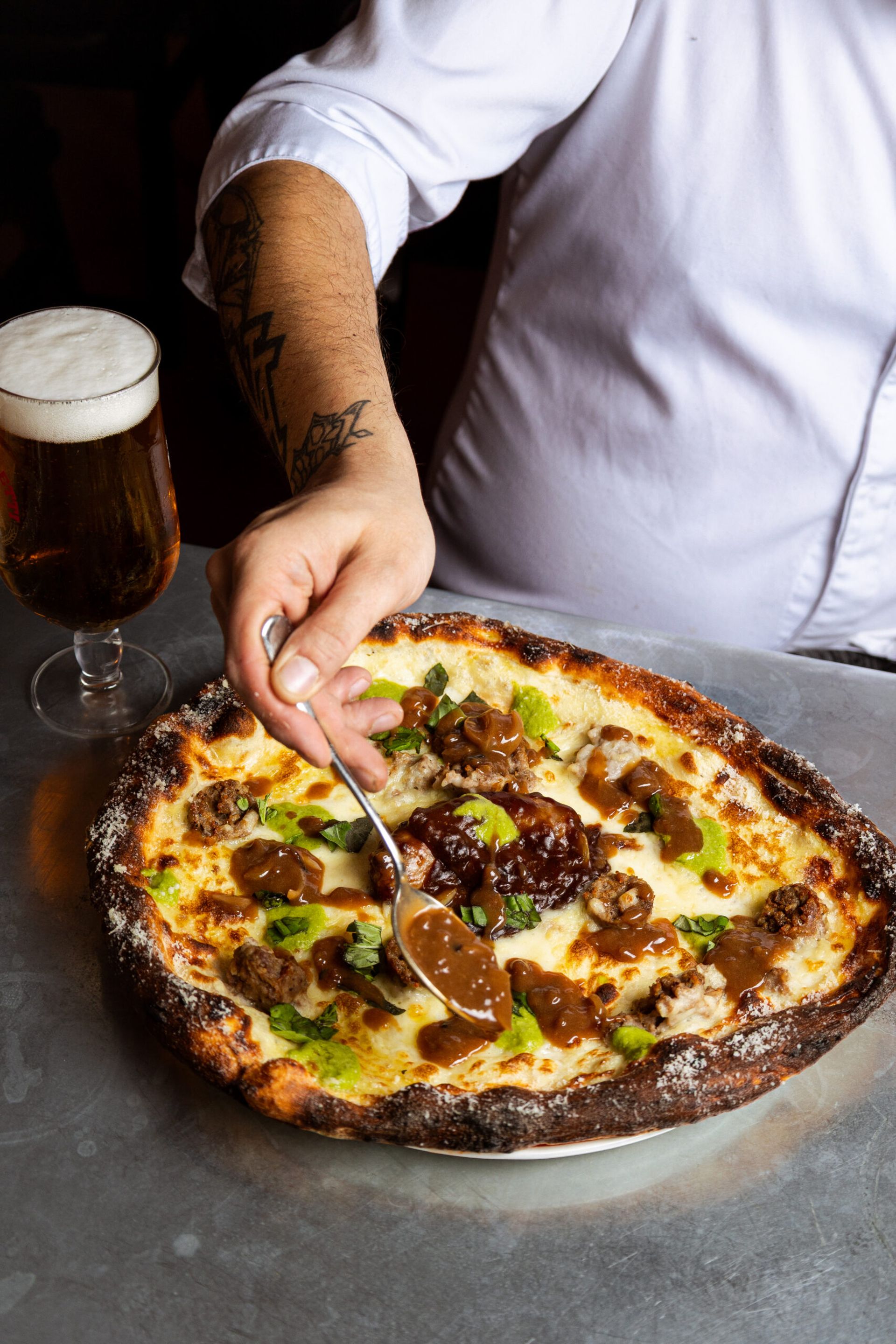 A person in a white chef’s jacket spoons brown sauce onto a freshly baked pizza with greens and cheese, while a glass of beer sits nearby on the grey table—perfect inspiration for your weekend guide.