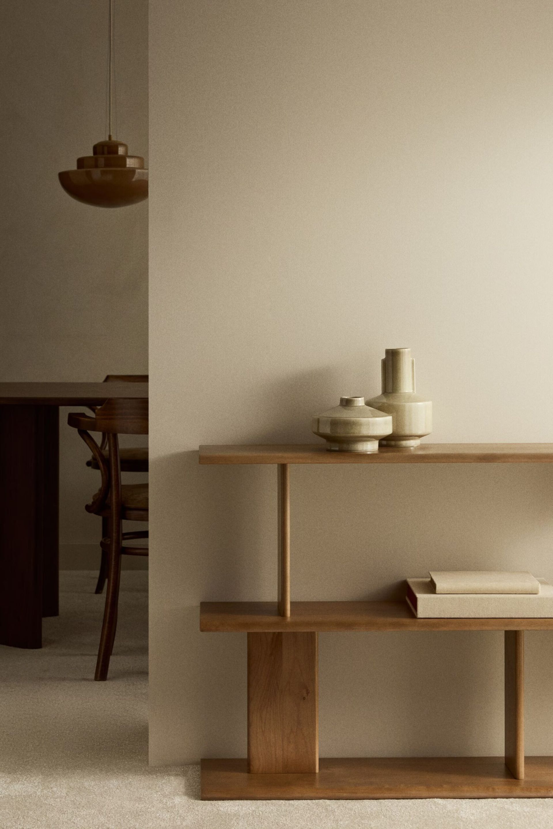 A minimalist interior featuring a wooden console table with neutral-toned vases and a closed book. High street homeware accents enhance the space, whilst a dining table, chairs, and pendant light are partially visible in the warm, muted palette.