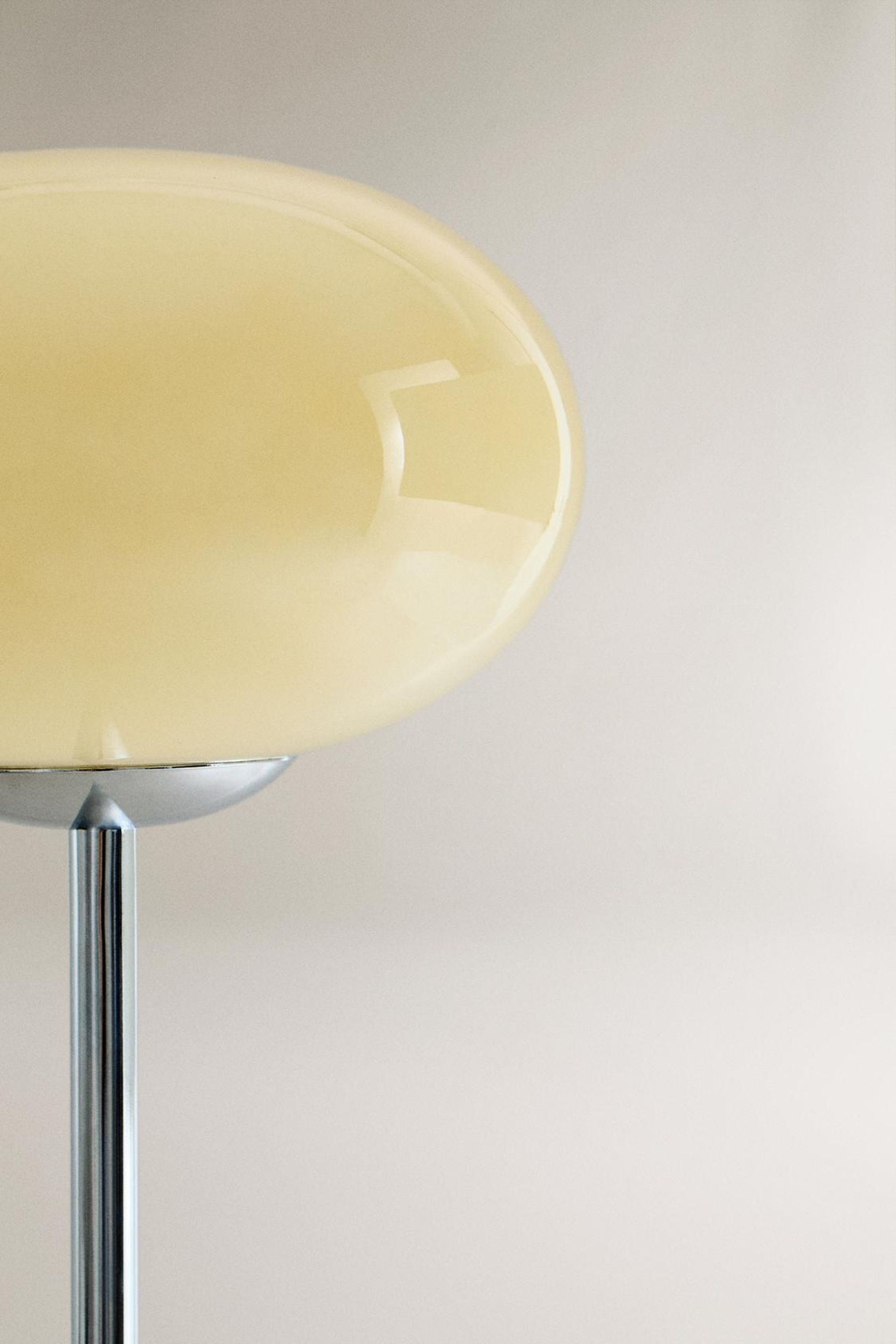 A close-up of a modern floor lamp, reminiscent of high street homeware, with a glossy, oval-shaped beige lampshade and a shiny metallic stand, set against a plain light-coloured background.