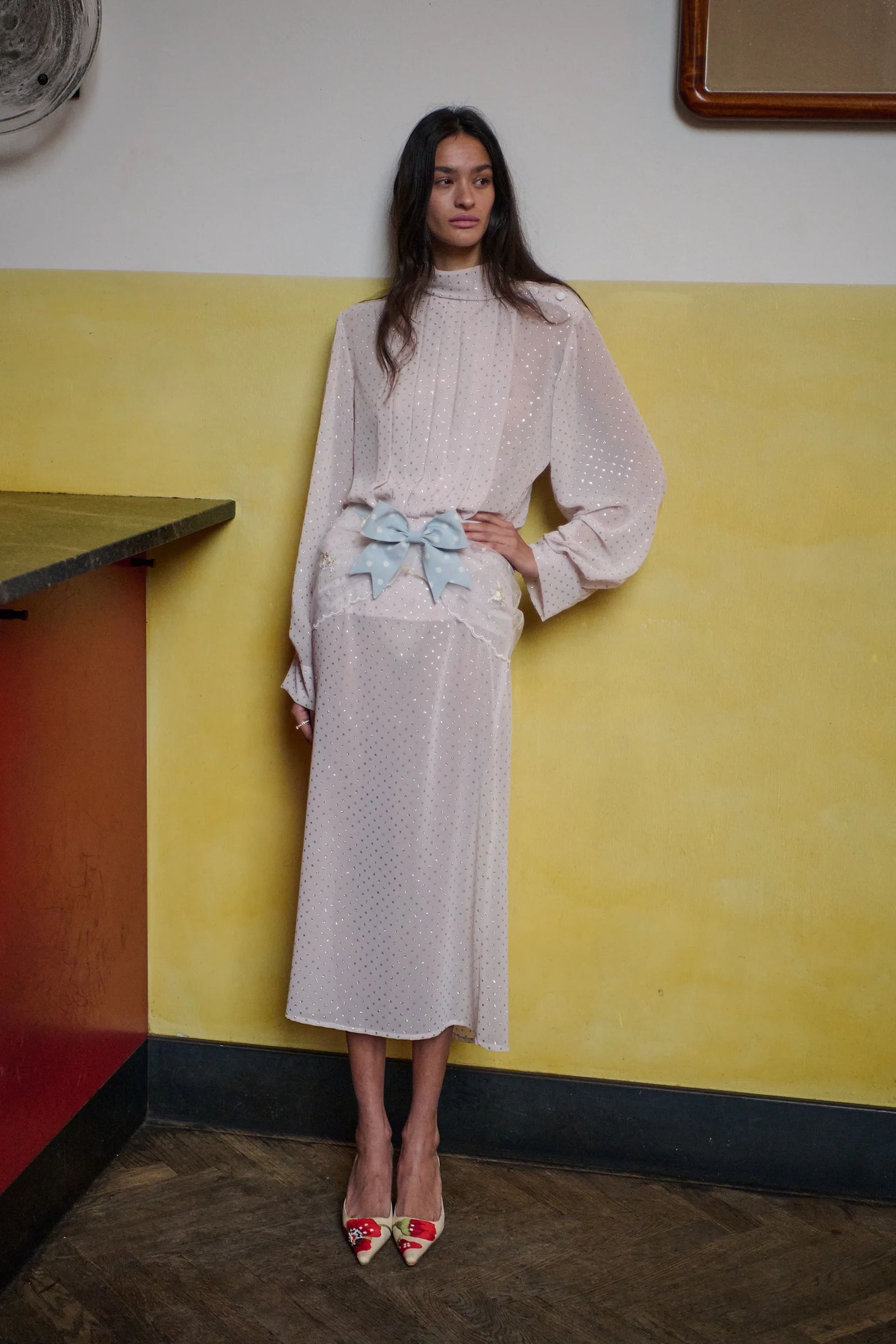 A woman in a sheer, pale pink midi dress with subtle polka dots, light blue bows at the waist, and floral pointed heels stands against a yellow and white wall—an elegant nod to spring/summer 2026 fashion trends.