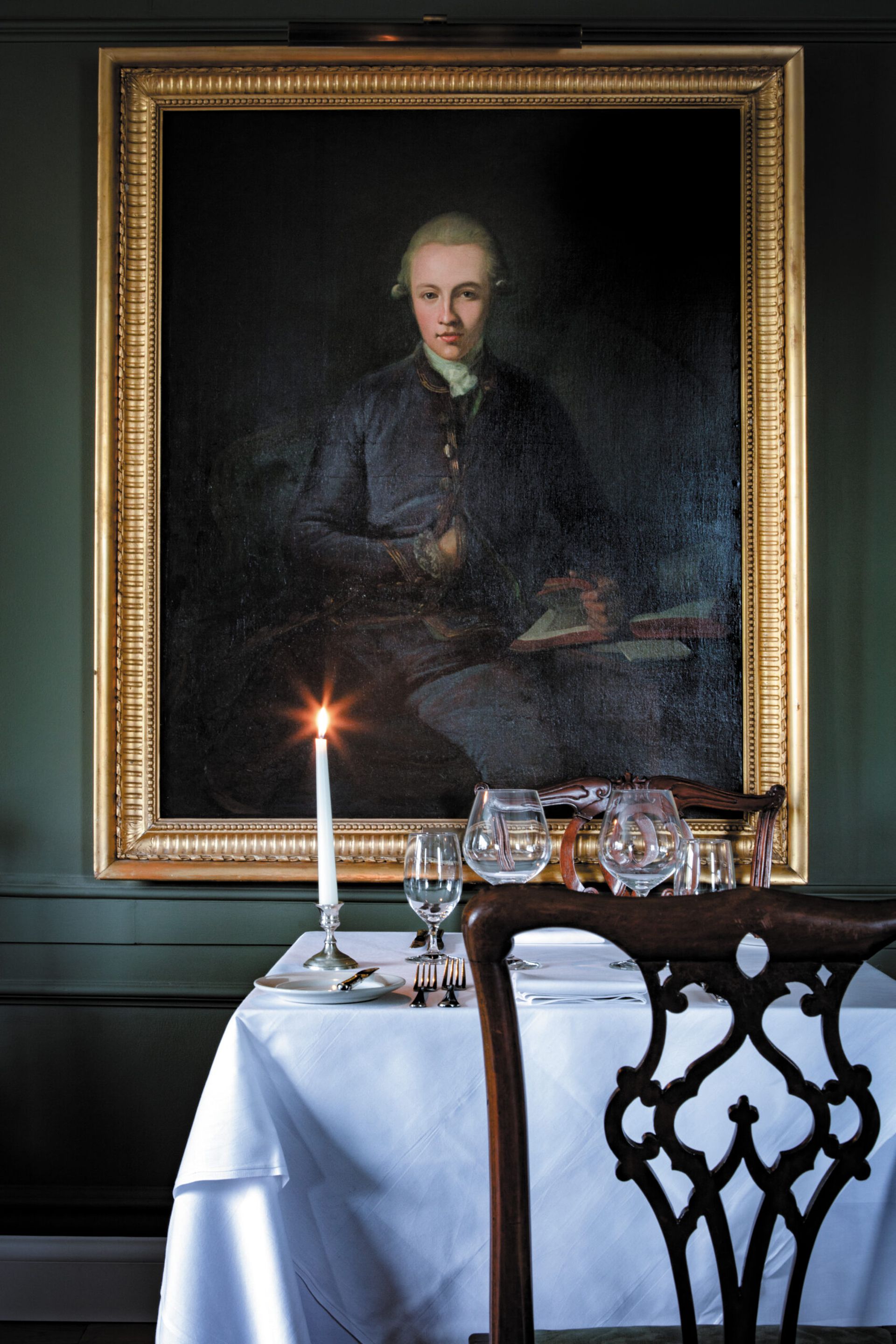 A candlelit dining table set with glasses and cutlery is positioned in front of a large, framed portrait of a person in historical clothing against a dark green wall at Kinloch Lodge.