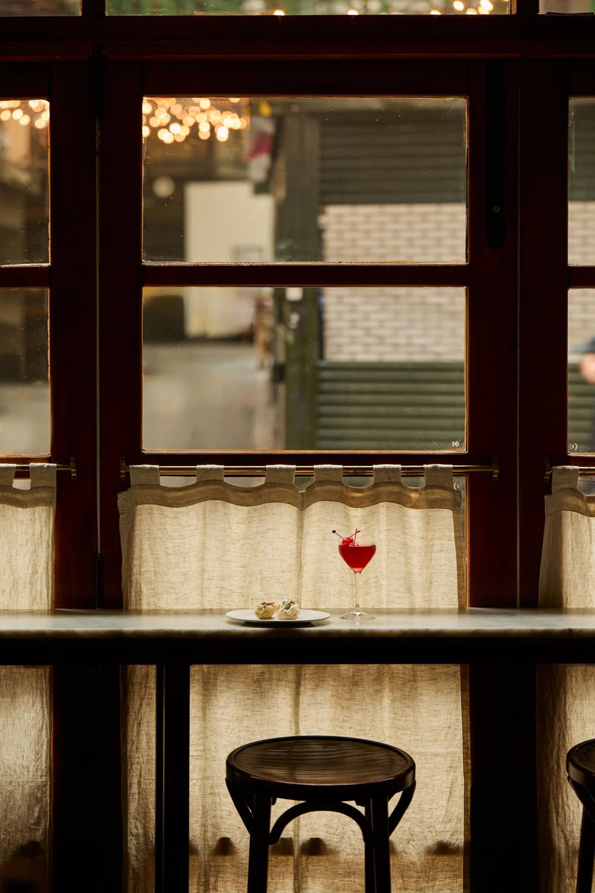 A dimly lit café interior with a high window, sheer curtains, and two empty stools at a bar. On the counter sits a cocktail—reminiscent of something Hannah Crosbie might recommend—and a small plate of food, blurred lights outside.
